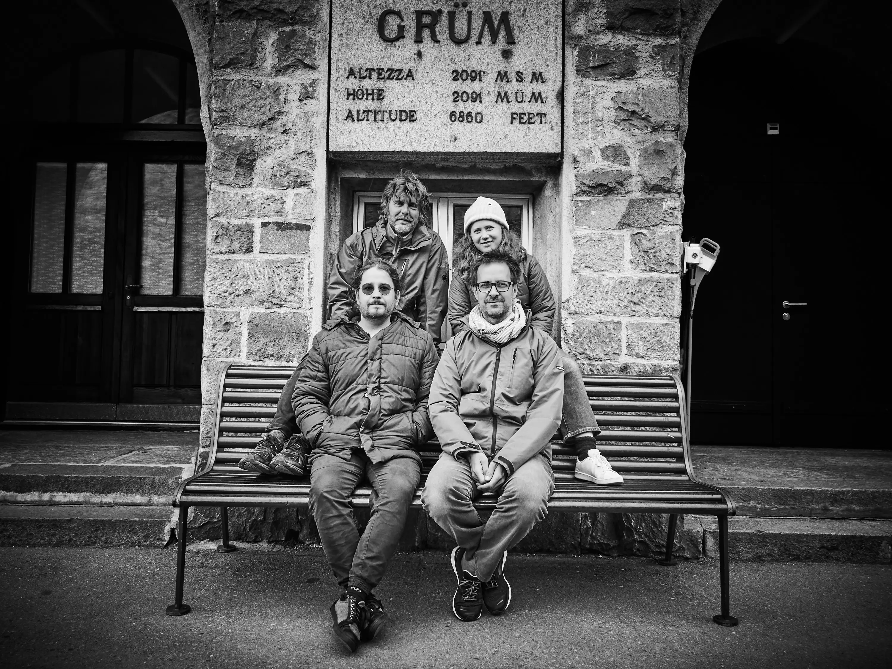 Quattro persone sorridenti posano su una panchina di fronte a un edificio in pietra con una scritta che indica l'altezza. Due sono sedute e due in piedi dietro. L'atmosfera è informale e accogliente.