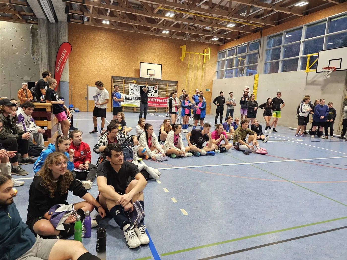 Un gruppo di persone sedute in una palestra, mentre assistono a un evento. Alcuni partecipanti sono in abbigliamento sportivo, e ci sono poster e attrezzature sportive sullo sfondo.