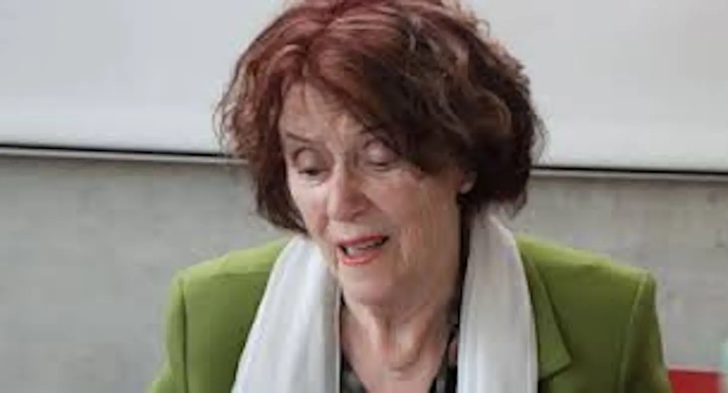 Donna anziana con capelli rossi ricci, indossa una giacca verde e una sciarpa bianca. È seduta e sembra assorta nella lettura o nella riflessione.