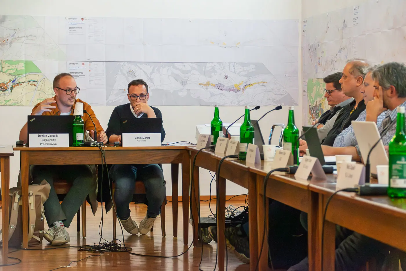 Un gruppo di persone sedute attorno a un tavolo durante una riunione, con mappe appese alle pareti. Due relatori sono di fronte a un computer portatile, mentre gli altri partecipanti ascoltano con bottiglie d'acqua verdi sul tavolo.