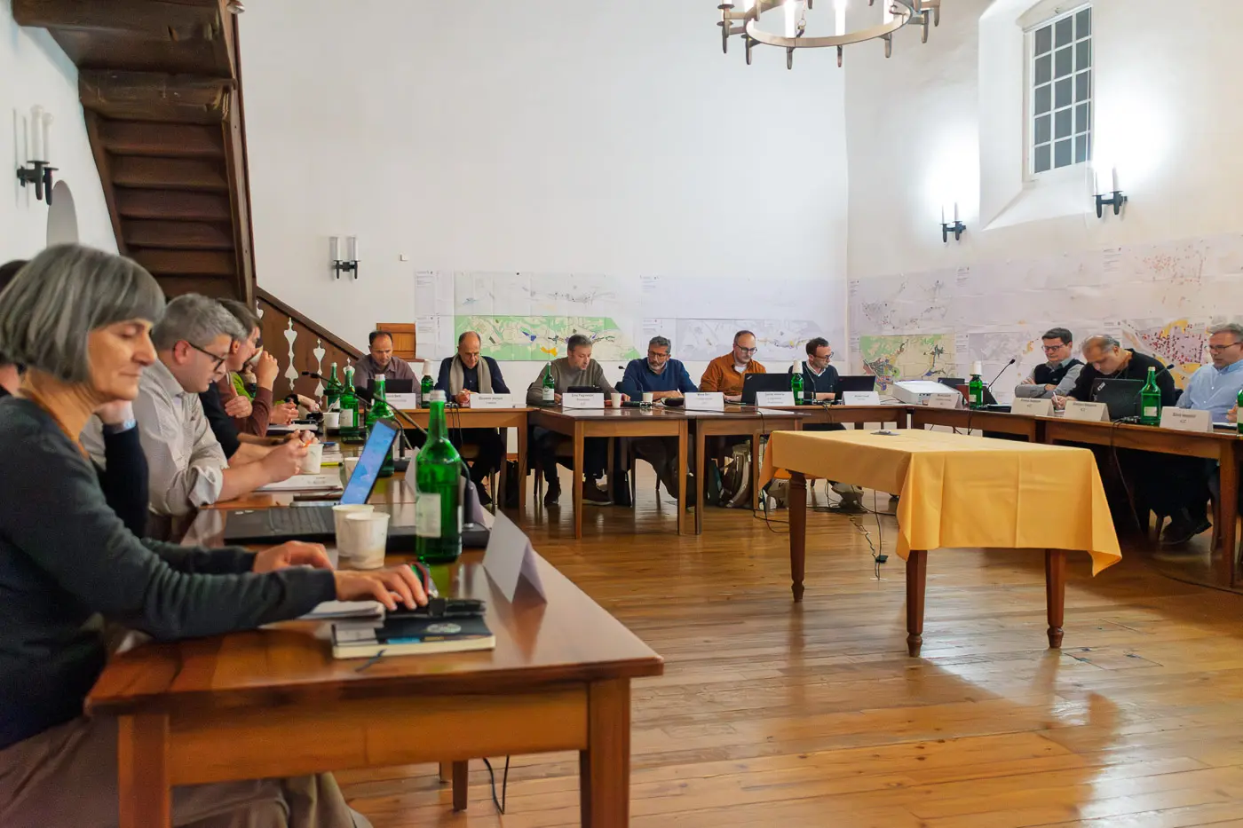 Un gruppo di persone sedute attorno a un grande tavolo in una sala di riunioni. Alcuni partecipanti sono concentrati su computer portatili, mentre altri discutono documenti e mappe appese alle pareti. Diverse bottiglie d'acqua sono sparse sui tavoli