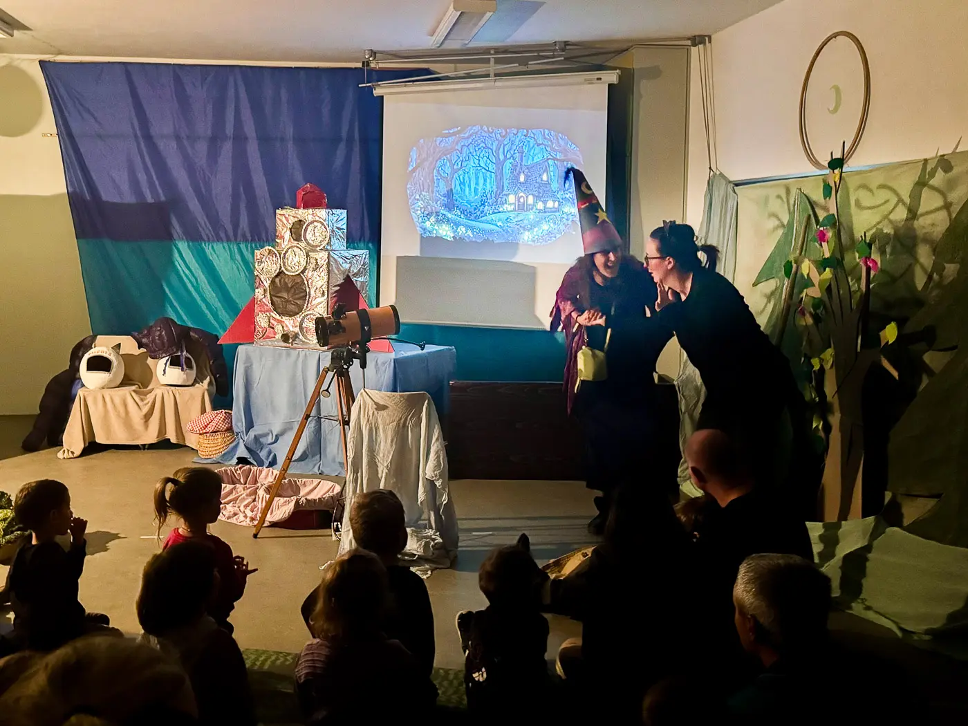 Una rappresentazione teatrale per bambini in corso, con attori vestiti in costume, un proiettore sullo sfondo che mostra un'immagine fantasy e un pubblico di bambini seduti.