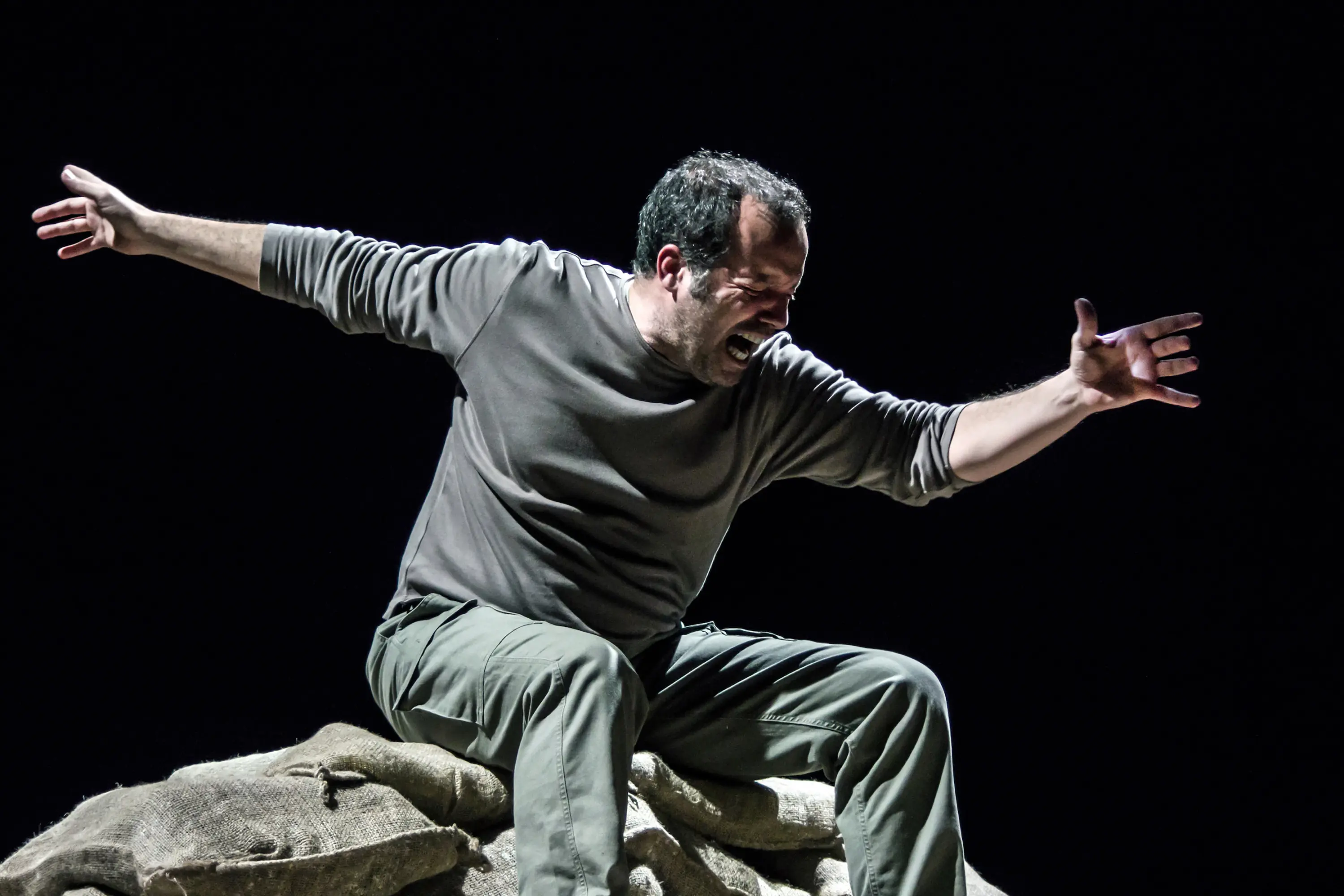 Un attore si esibisce su un palco buio, seduto su un cumulo di sacchi, mentre esprime intensa emozione con gesti ampi e un'espressione facciale coinvolgente.