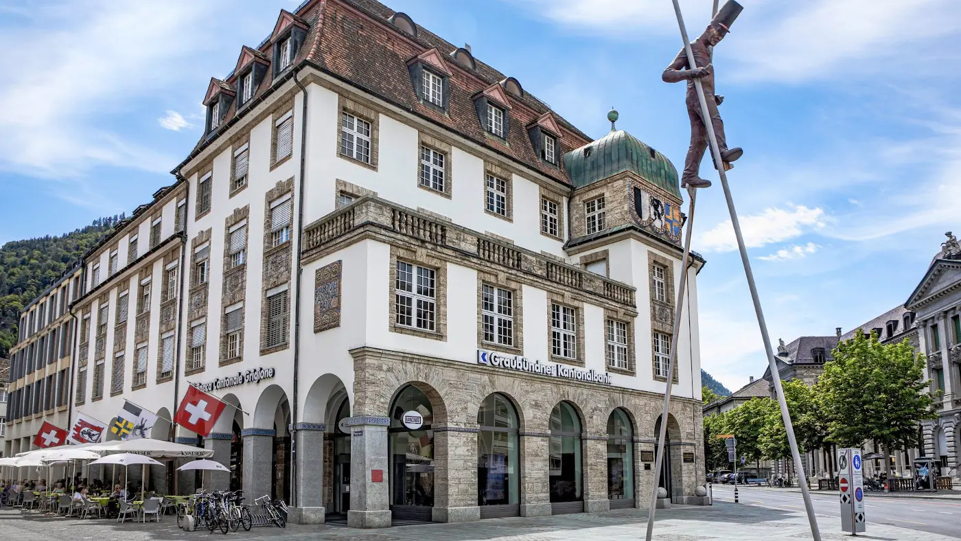 L'edificio storico con facciata bianca e dettagli in pietra si trova in una piazza. Sul tetto è presente una cupola verde. Bandiera svizzera e altre bandiere decorano l'entrata. Una statua di un uomo stilizzato è visibile acc
