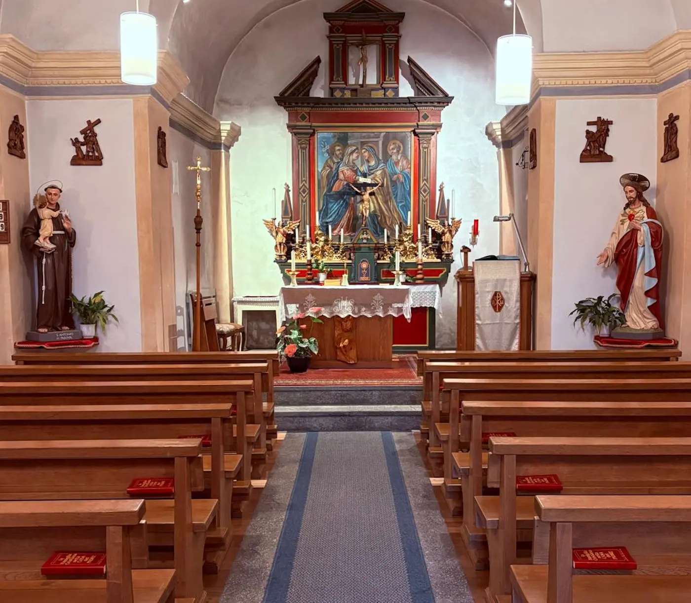 Interno di una chiesa con un altare centrale decorato da un dipinto. Ai lati dell'altare ci sono statue di santi. Panchine in legno disposte in fila e un tappeto che conduce all'altare. Illuminazione soffusa e atmosfera tranquilla.