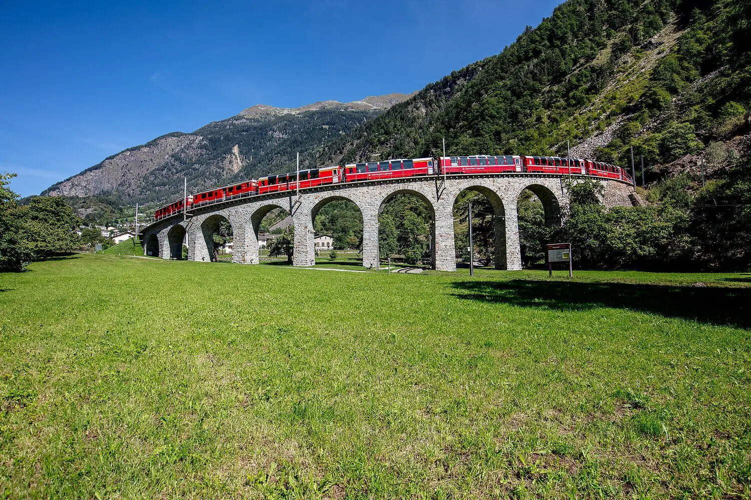 Un treno rosso attraversa un ponte ad archi in pietra, immerso in un paesaggio verdeggiante con montagne sullo sfondo. Il cielo è sereno e blu.