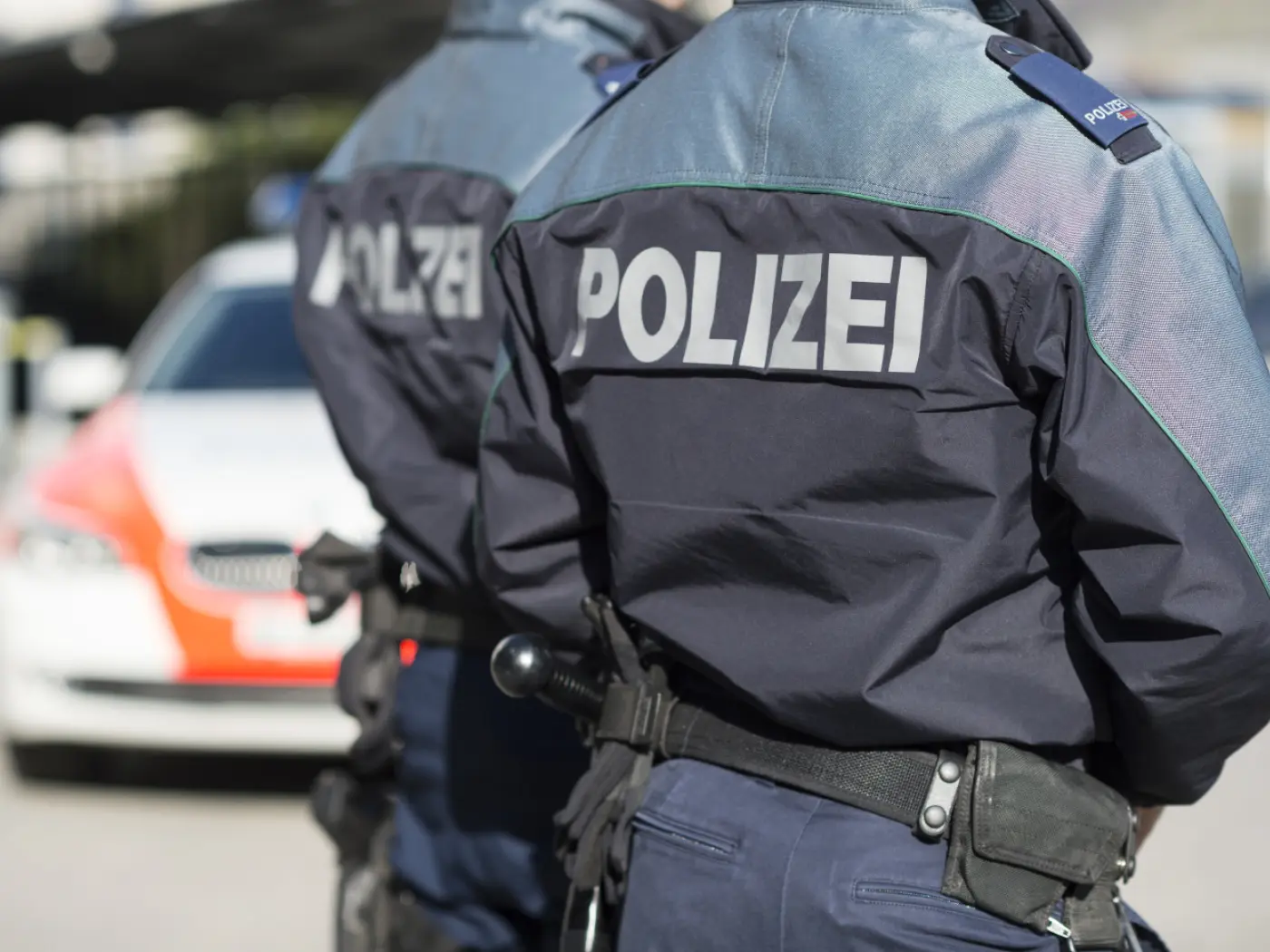 Due agenti di polizia in uniforme con la scritta "POLIZEI" sulle spalle, in primo piano. Sullo sfondo si vede un'auto della polizia.