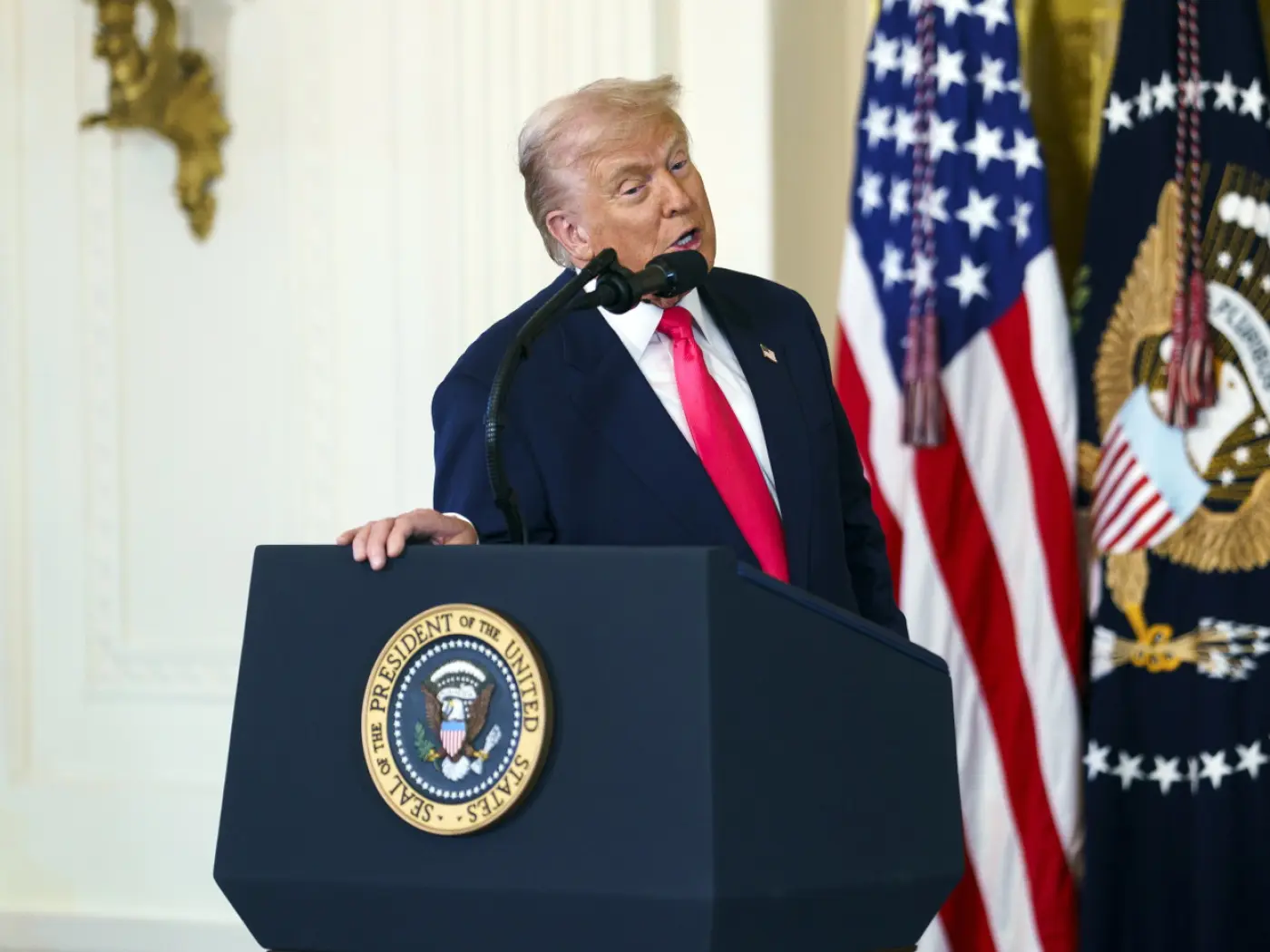 Uomo in giacca e cravatta rossa parla a un podio con il sigillo presidenziale, accanto a bandiere americane.