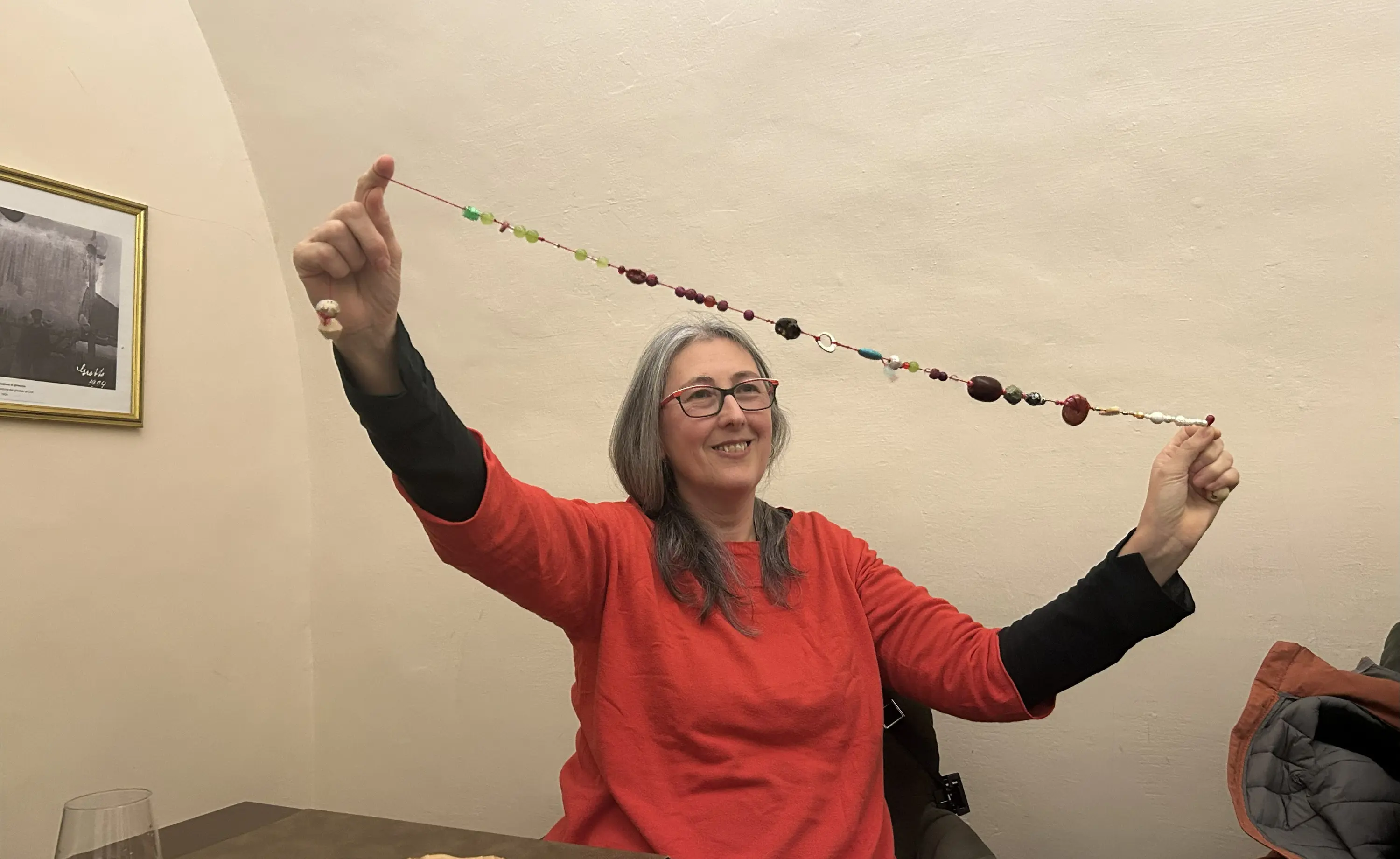 Donna sorridente con capelli lunghi e grigi, che tiene in alto una lunga collana colorata. Indossa una maglietta rossa e si trova all'interno di un ambiente illuminato.