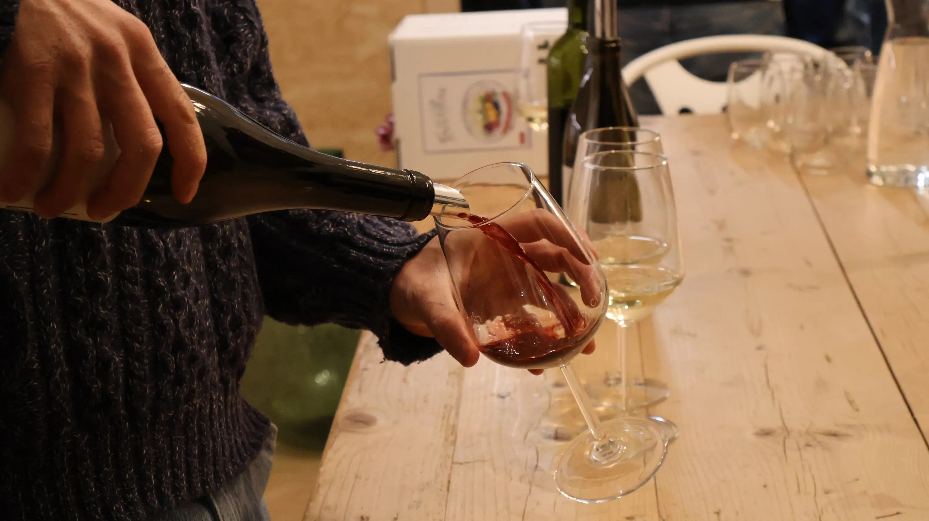 Un uomo versa del vino rosso in un bicchiere su un tavolo di legno. Sul tavolo ci sono anche altri bicchieri e una bottiglia di vino.