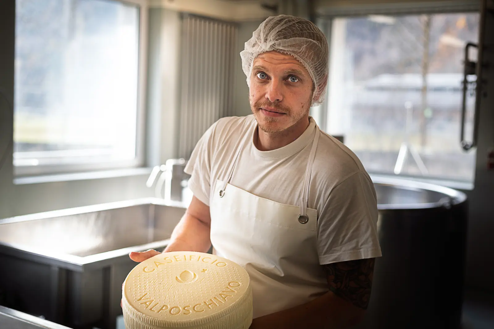 Un casaro in un laboratorio di produzione di formaggio tiene in mano un grande formaggio rotondo, con il logo "Caseificio Valposchiano" visibile in alto. Indossa un grembiule bianco e una cuffia per capelli, con attrezzature per la produzione