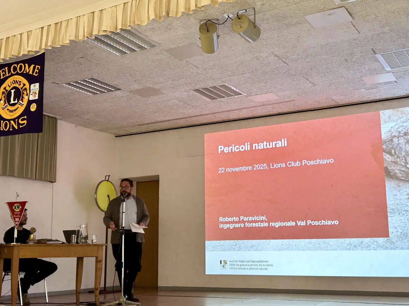 Un uomo in piedi parla a un pubblico durante una presentazione. Sullo schermo dietro di lui è visibile il titolo "Pericoli naturali" con la data e il nome del relatore. Un altro uomo è seduto davanti, probabilmente come pubblico. I loghi del