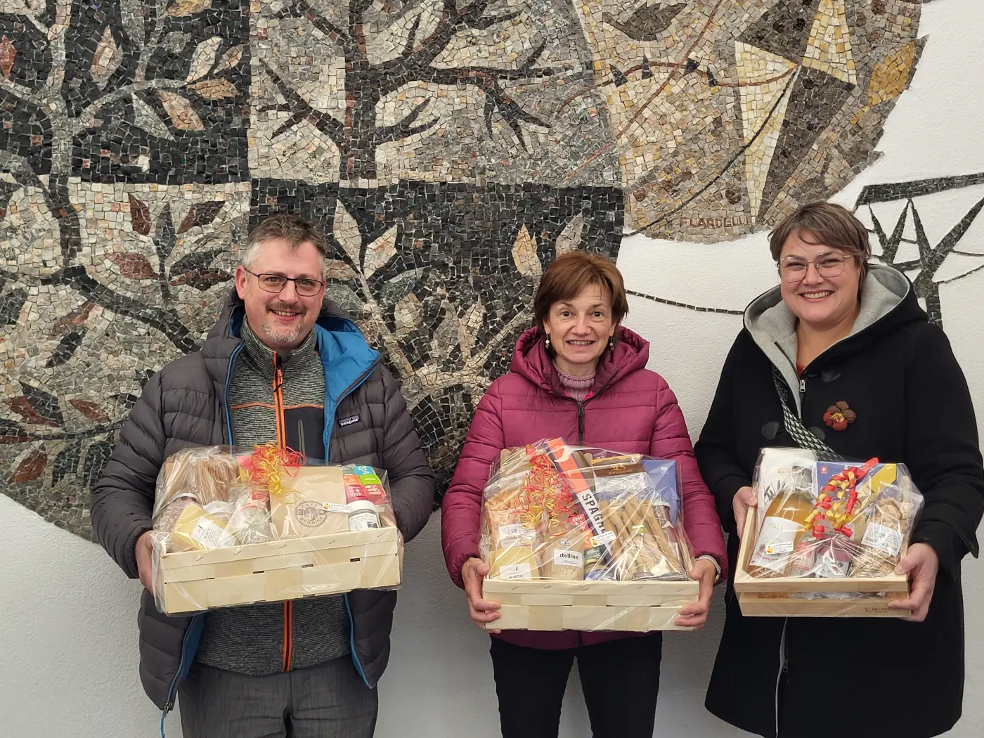 Tre persone sorridenti tengono in mano cesti di prodotti gastronomici. Sullo sfondo, un mural decorativo con un albero rappresentato in stile mosaico.