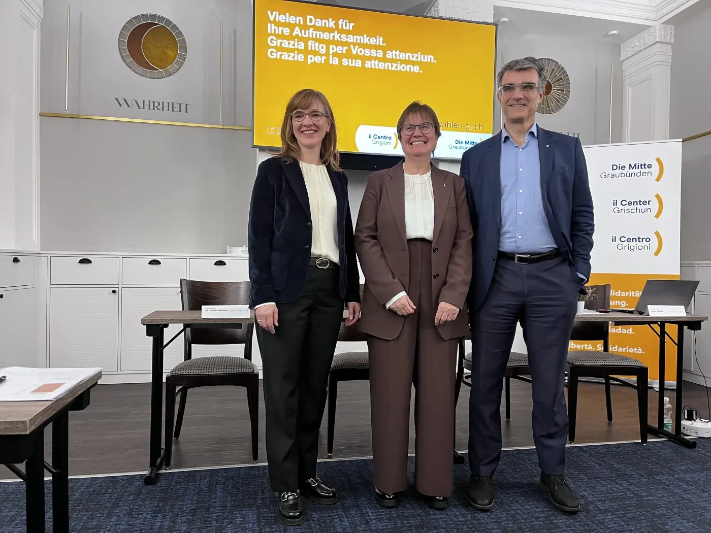 Tre persone sorridenti posano insieme in una sala conferenze. Sullo sfondo, uno schermo mostra un messaggio di ringraziamento in tedesco e italiano. Il contesto appare formale.