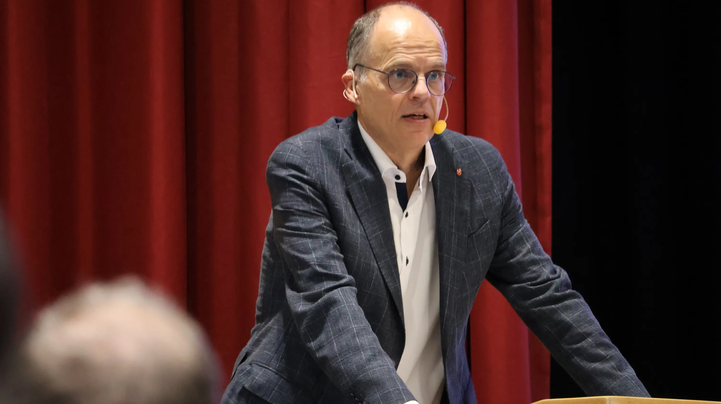 Uomo in giacca a quadri che parla da un podio con un microfono, di fronte a un pubblico, su sfondo rosso.