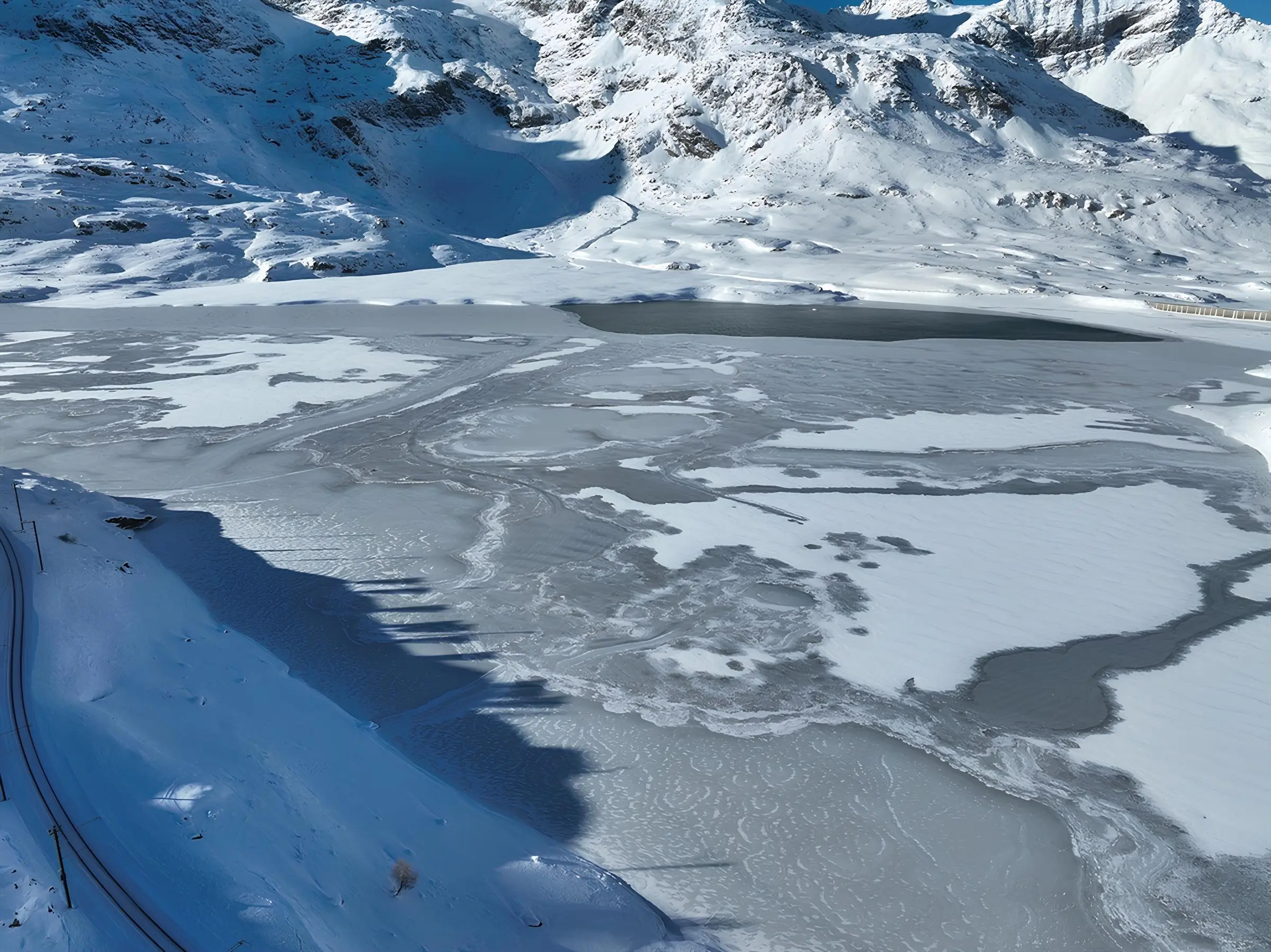 Paesaggio montano innevato con un lago ghiacciato. Le cime delle montagne sono coperte di neve e il sole illumina la scena, creando ombre sul ghiaccio