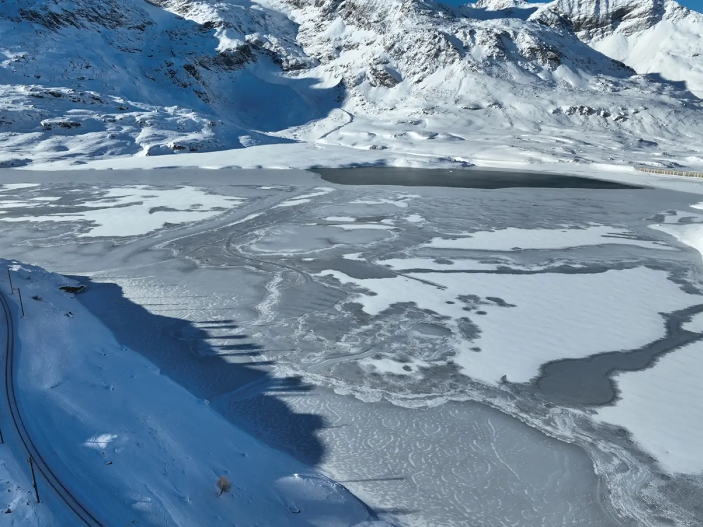 Paesaggio invernale con un lago ghiacciato circondato da montagne nunate. La superficie del lago presenta disegni ghiacciati, mentre il cielo è sereno.