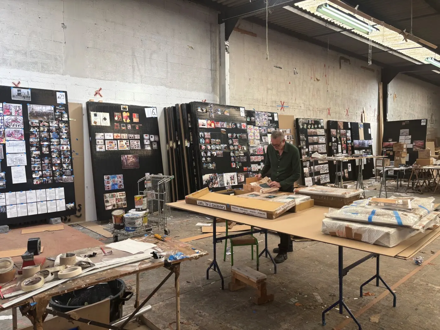Un uomo sta lavorando in un laboratorio spazioso, circondato da pareti ricoperte di fotografie e appunti. Tavoli di lavoro e materiali artistici sono sparsi nel locale,