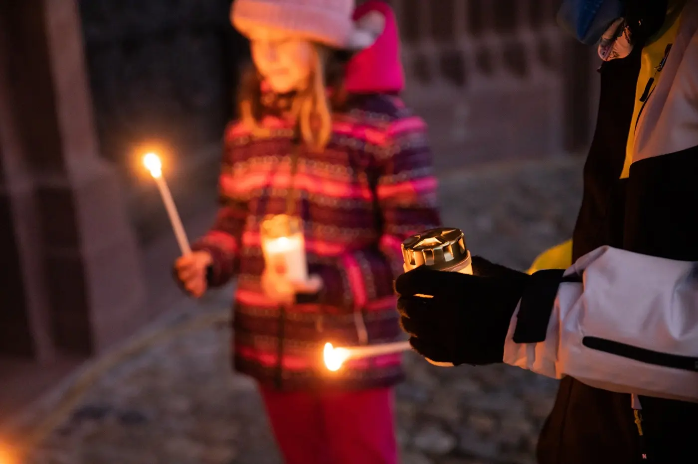 Due bambini partecipano a un evento serale, tenendo candele accese e lanterne. Indossano giacche pesanti, guanti e cappelli per il freddo