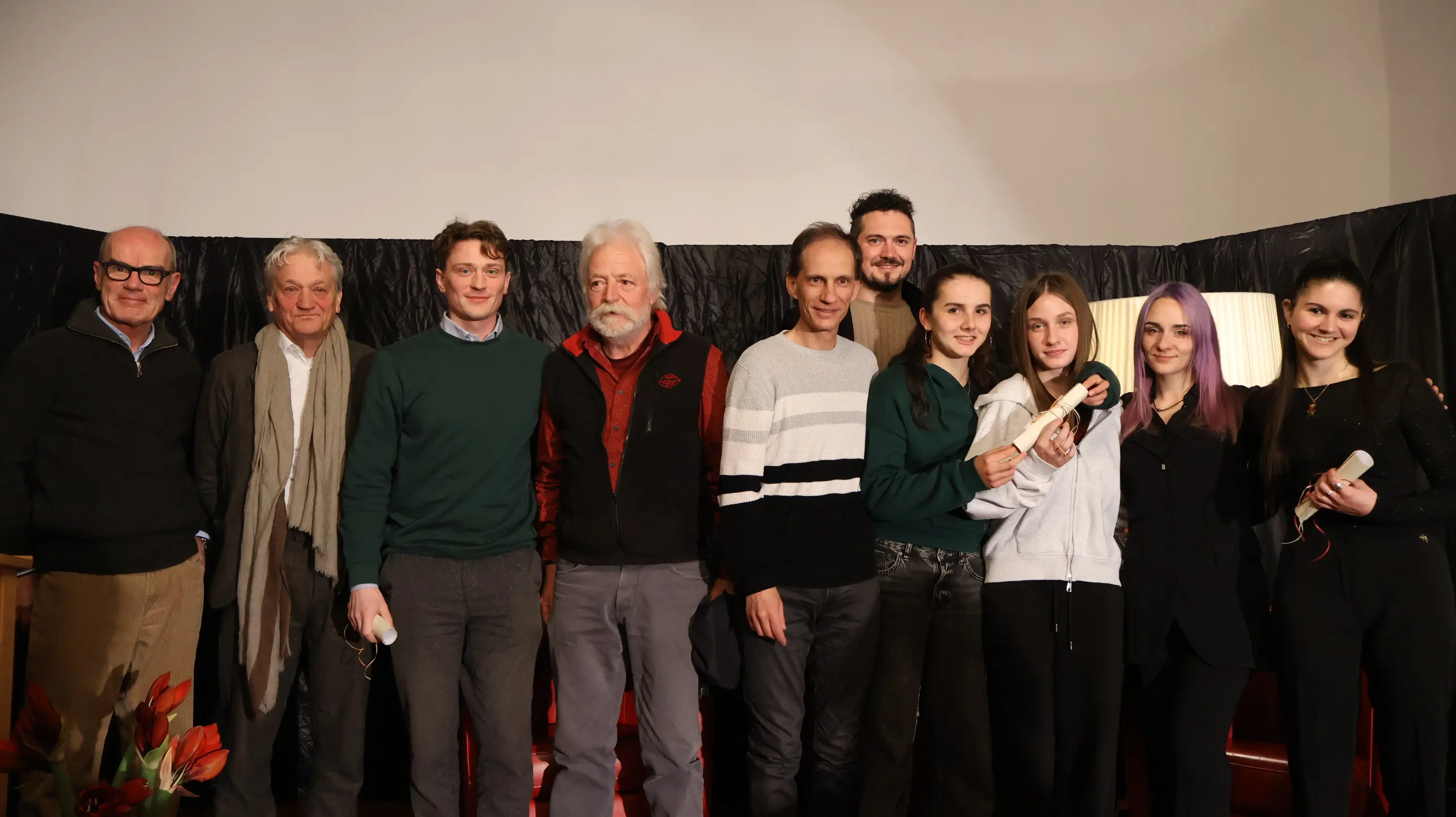 Gruppo di otto persone sorridenti, in abiti casual, su un palco con uno sfondo scuro. Alcuni hanno oggetti in mano, mentre hanno pose amichevoli e gio