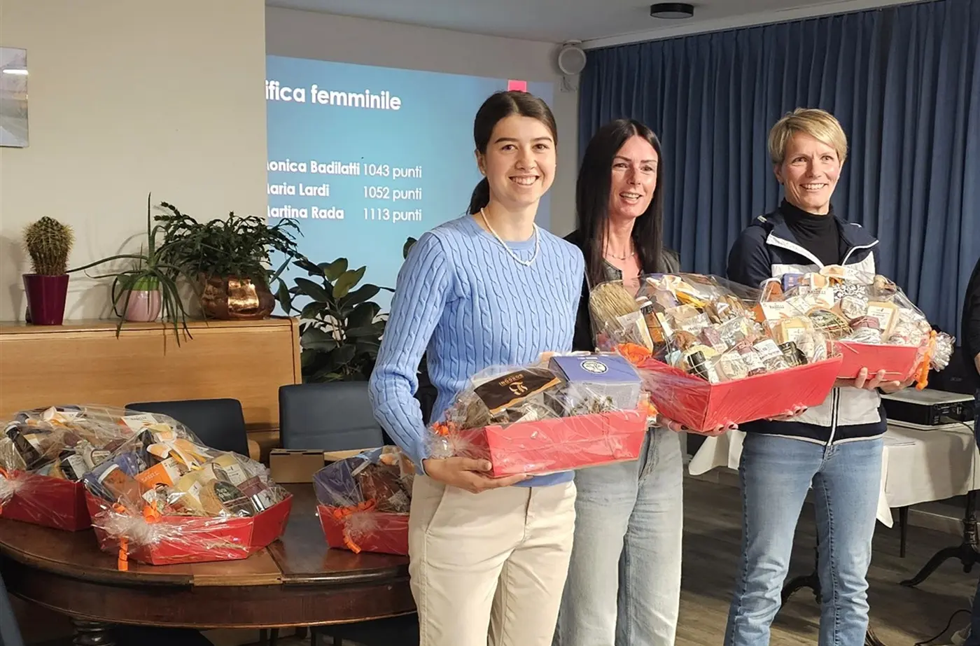 Tre donne sorridenti tengono in mano grandi cesti di prodotti dolciari. Sullo sfondo, una presentazione e alcune piante in vaso.