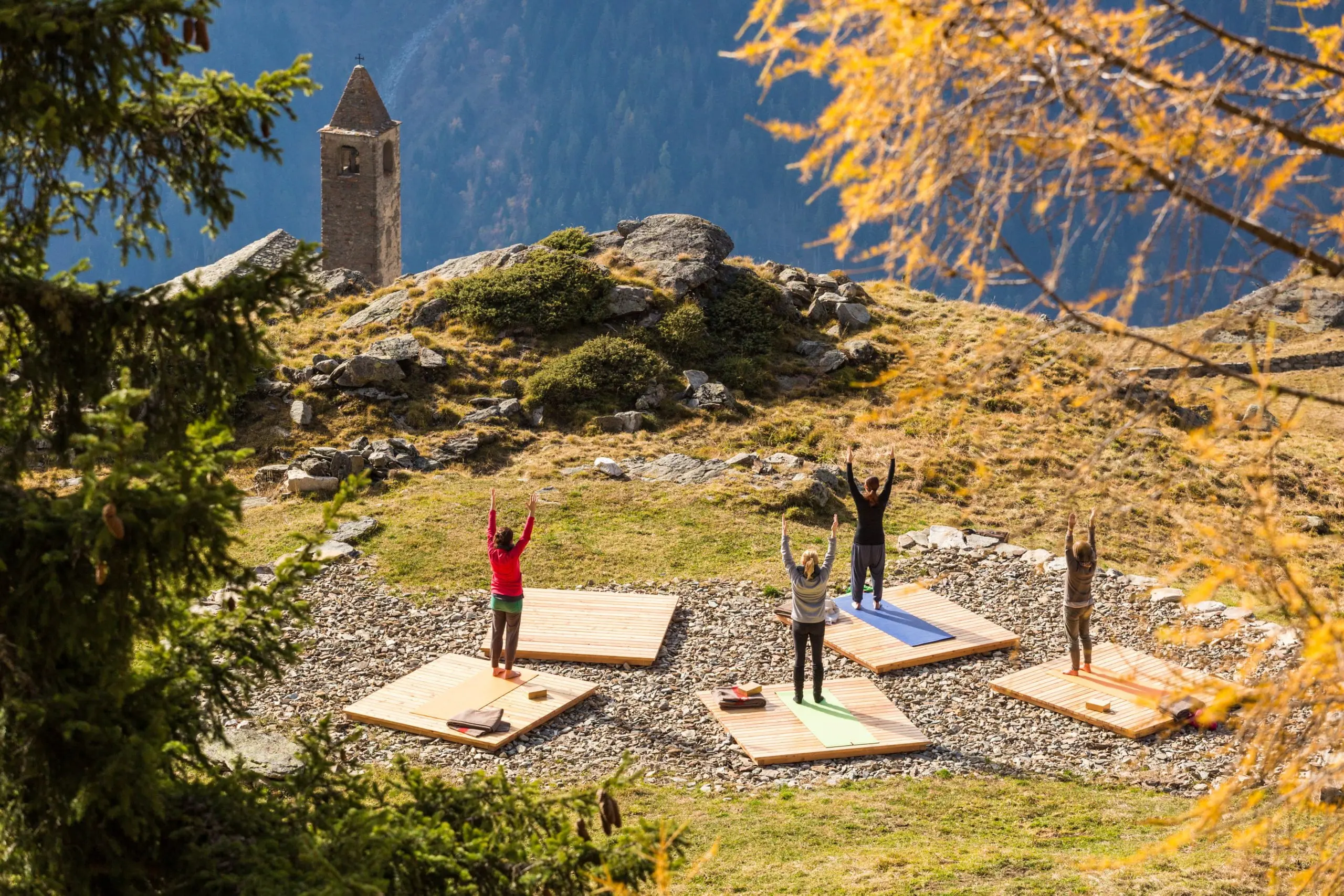 Gruppo di persone che praticano yoga all'aperto su tappetini, circondati da alberi e montagne in autunno. Una torre campanaria si erge sullo sf