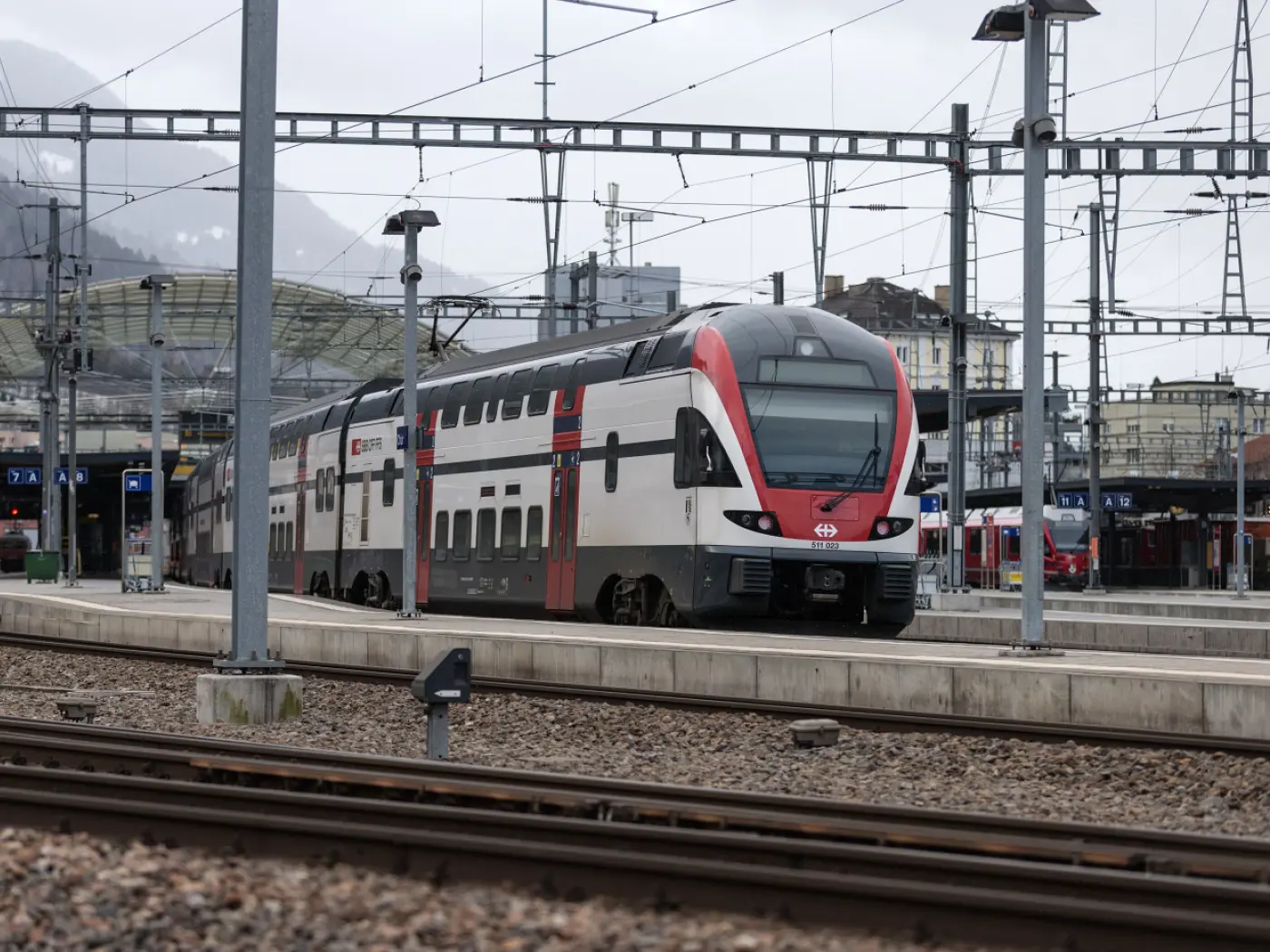 Un treno moderno con una livrea rossa e bianca si ferma su una piattaforma di una stazione ferroviaria. Sullo sfondo si vedono altre linee ferroviarie e