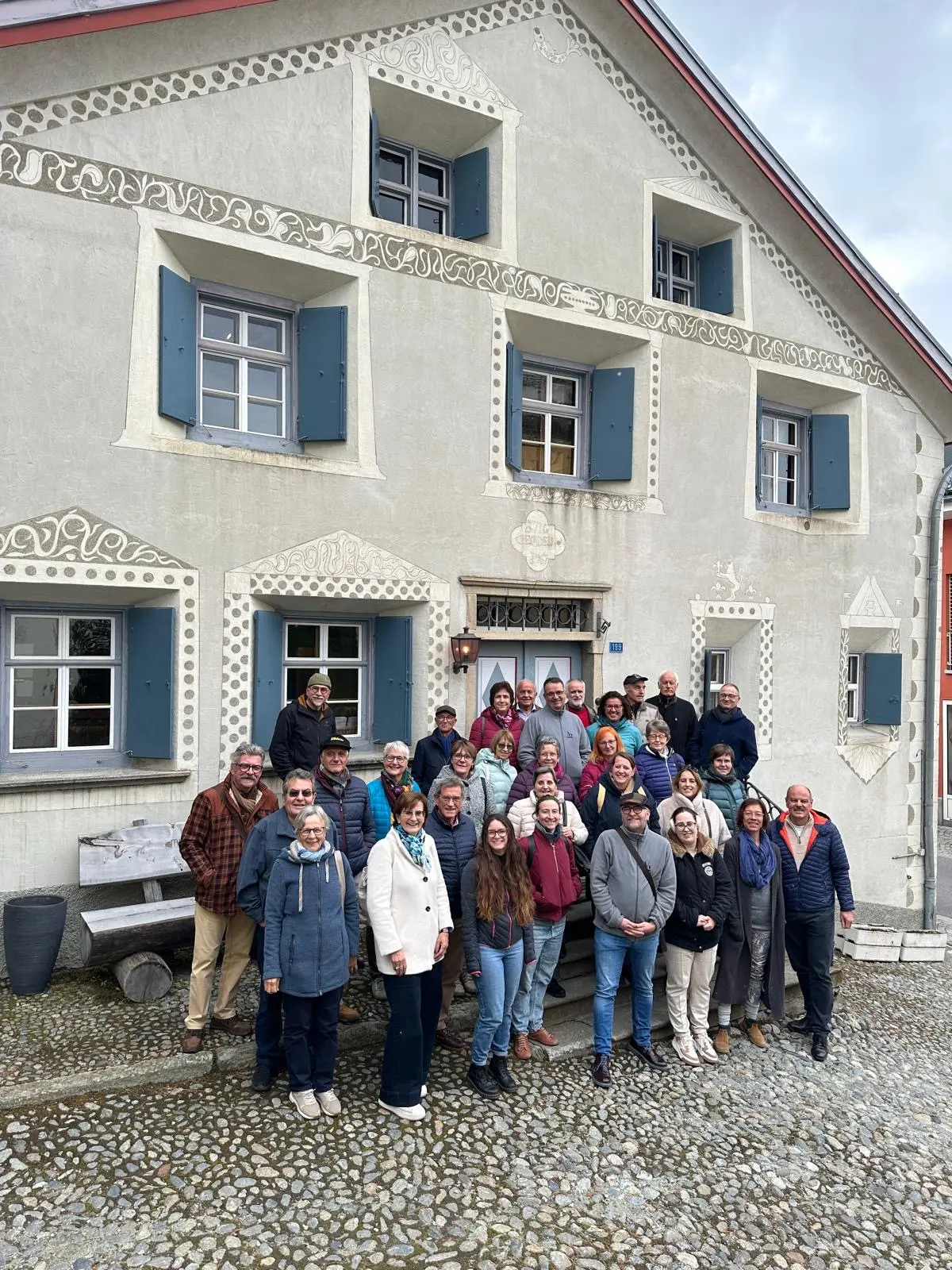 Gruppo di persone riunite di fronte a un edificio storico con finestre azzurre e decorazioni artistiche. Si vedono sorrisi e vari stili di abbigliamento,