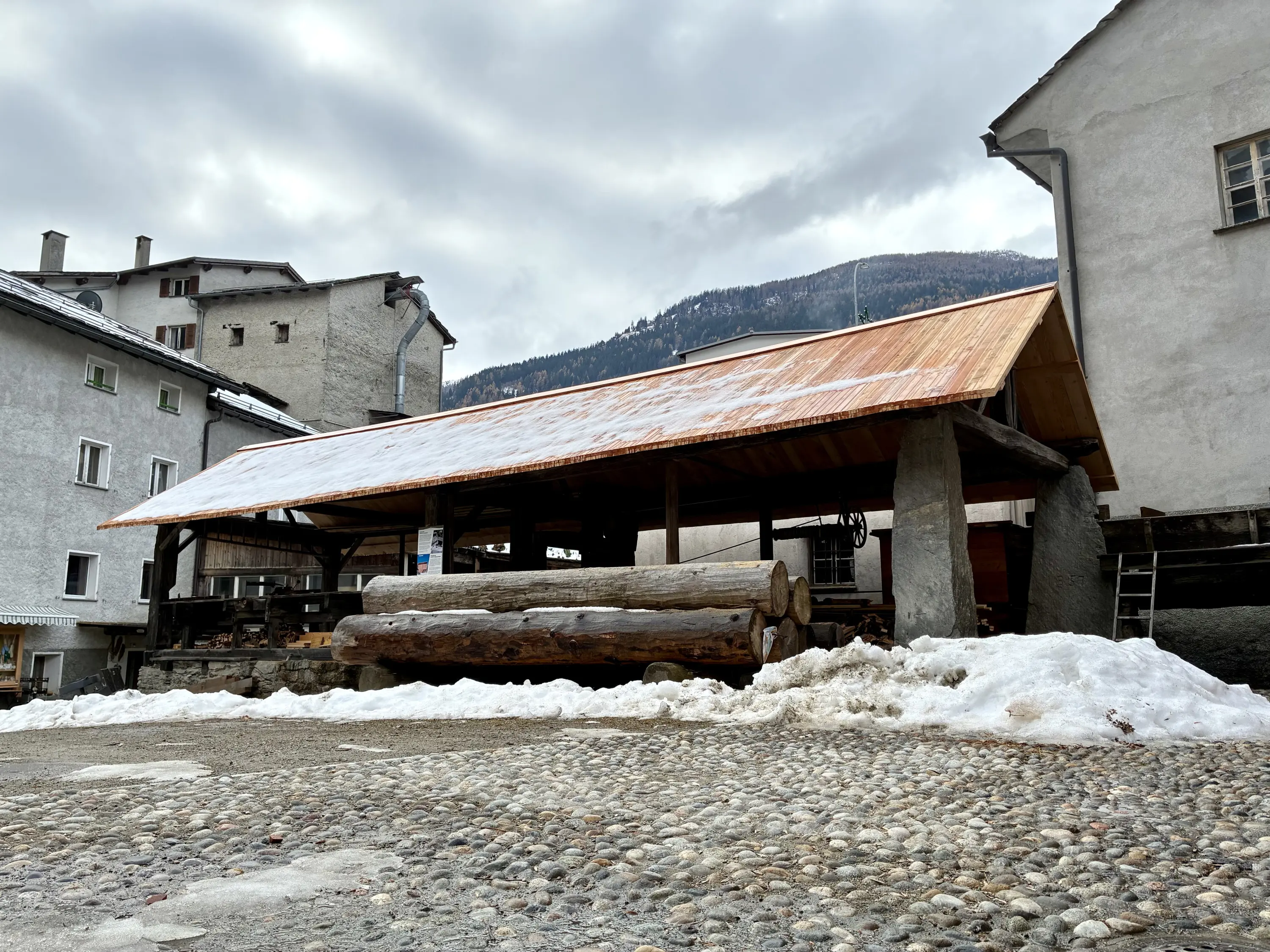 Un edificio tradizionale con tetto in metallo, situato in una piazza. Si vedono tronchi di legno disposti all'esterno e montagne sullo sfondo, con