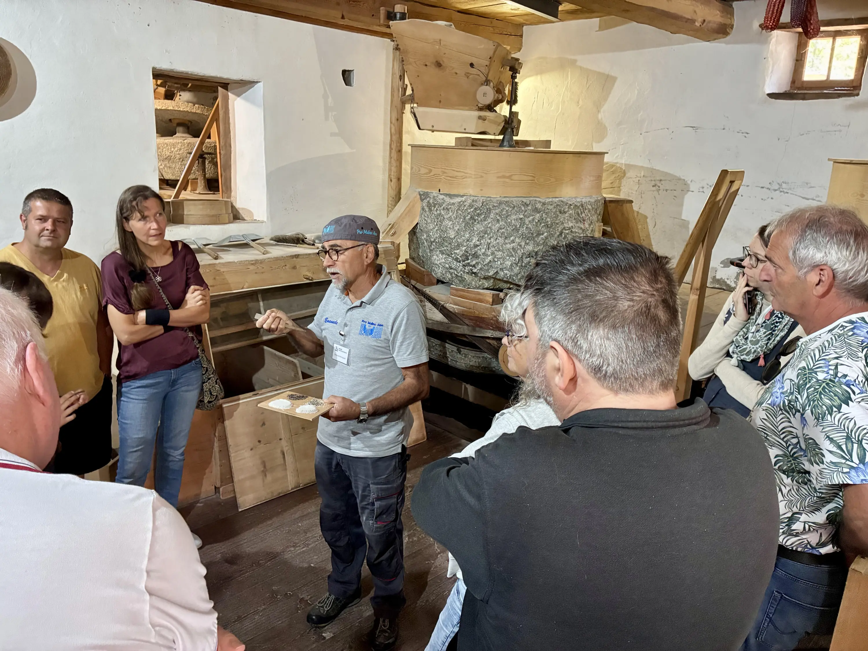 Un gruppo di persone ascolta attentamente un uomo che spiega qualcosa in un ambiente rustico, probabilmente all'interno di un'antica fabbrica o mulino. La stanza è caratterizzata