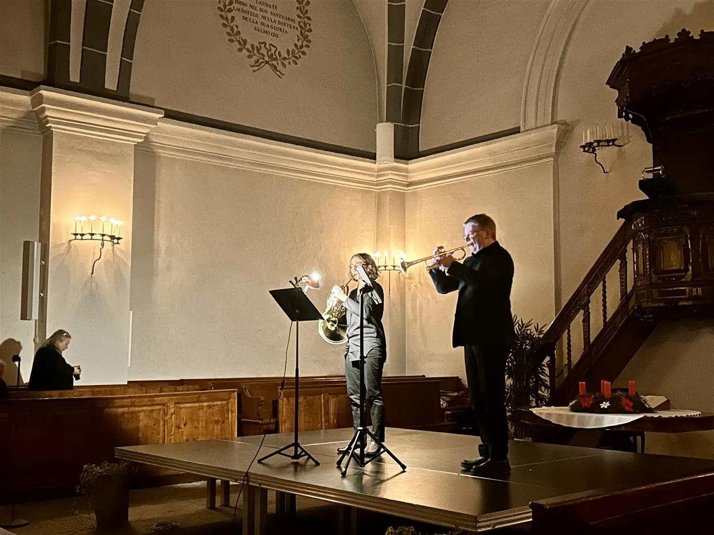 Due musicisti si esibiscono in una chiesa. Uno suona il trombone, mentre l'altro suona la tromba. Sullo sfondo, una scalinata in leg