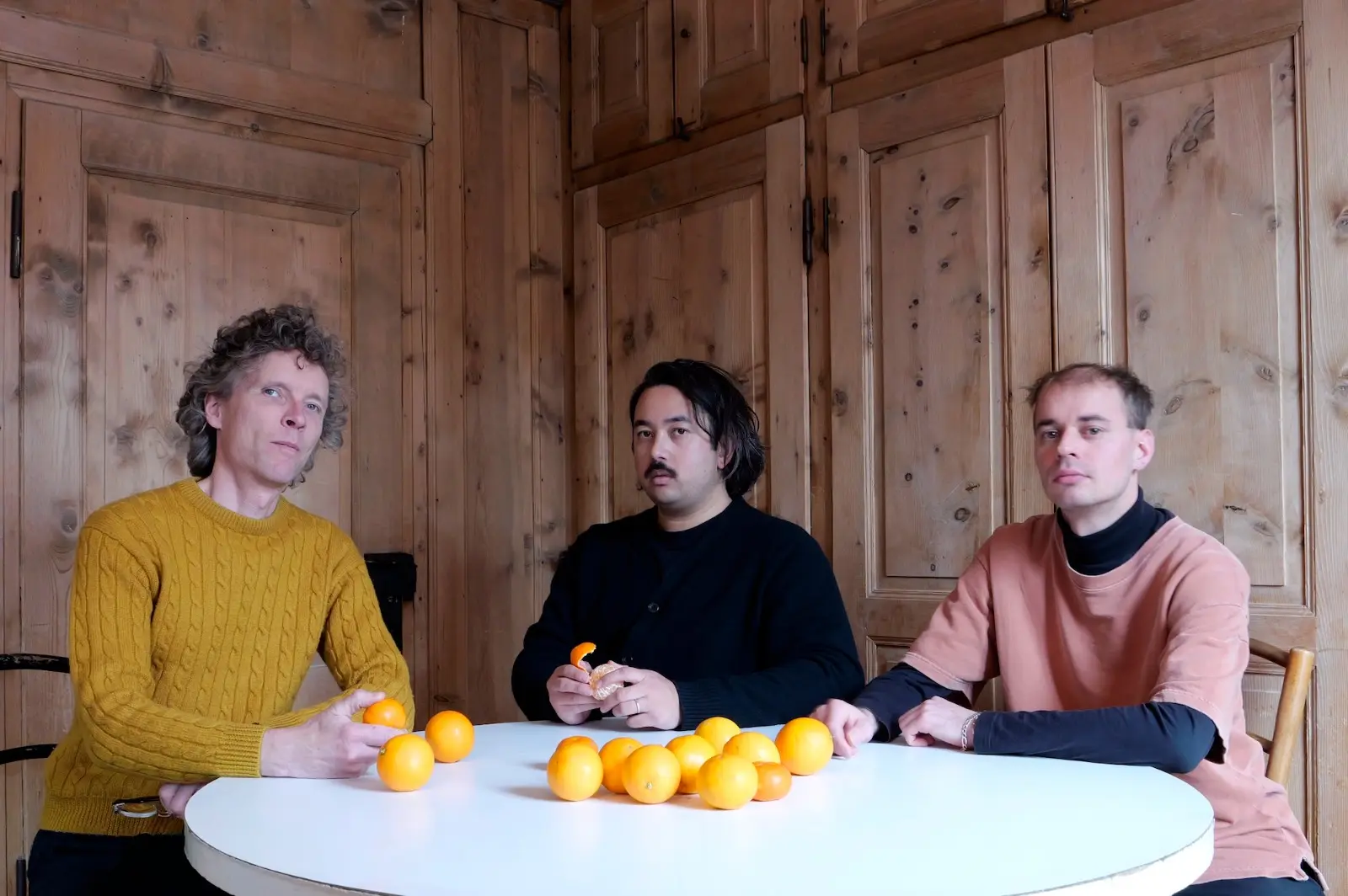 Tre uomini seduti attorno a un tavolo bianco. Hanno diversi tipi di maglioni. Sul tavolo ci sono arance disposte in modo casuale. Sullo sfondo