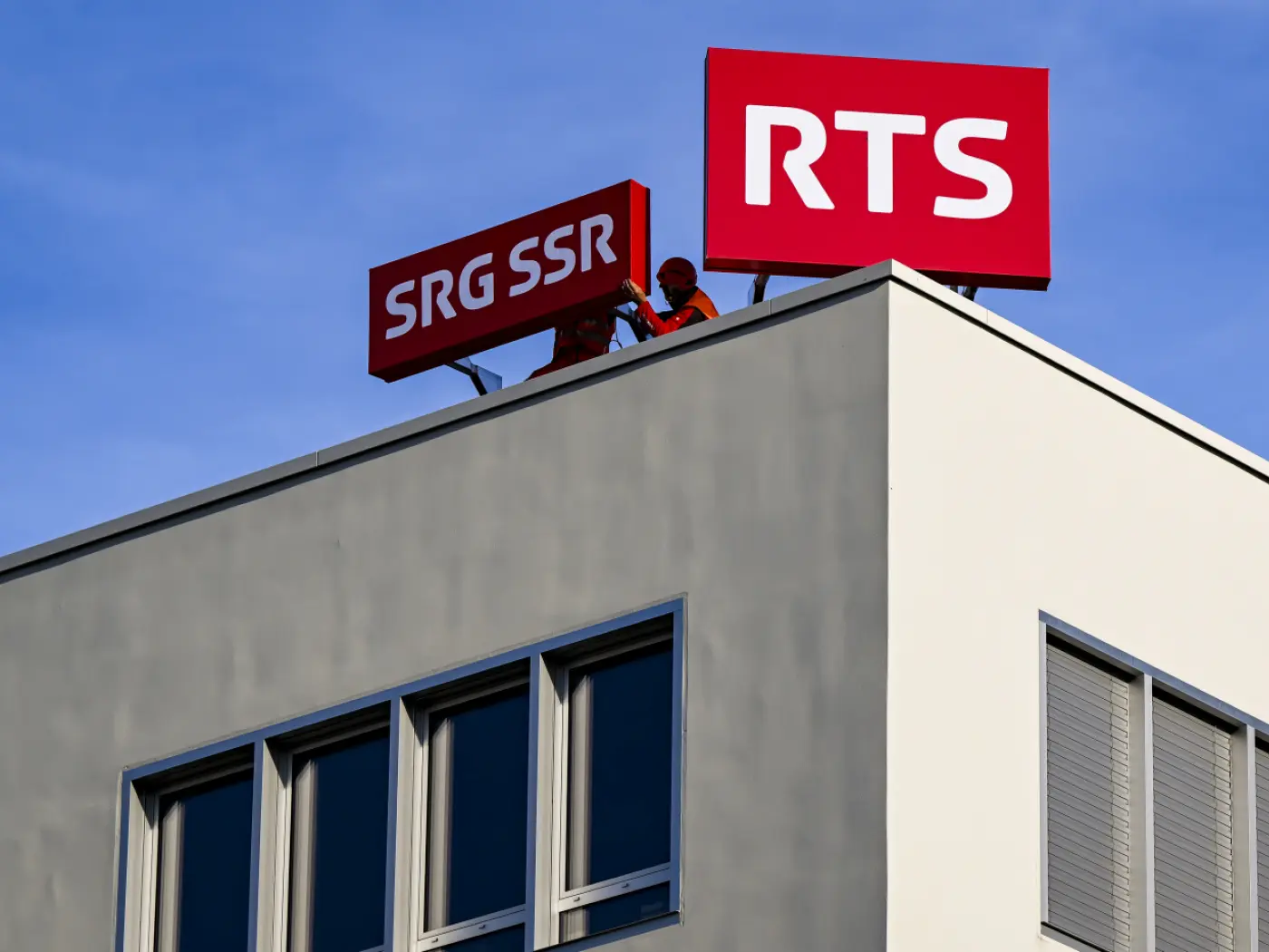 Due operai stanno installando segnali di SRG SSR e RTS sul tetto di un edificio moderno sotto un cielo azzurro.