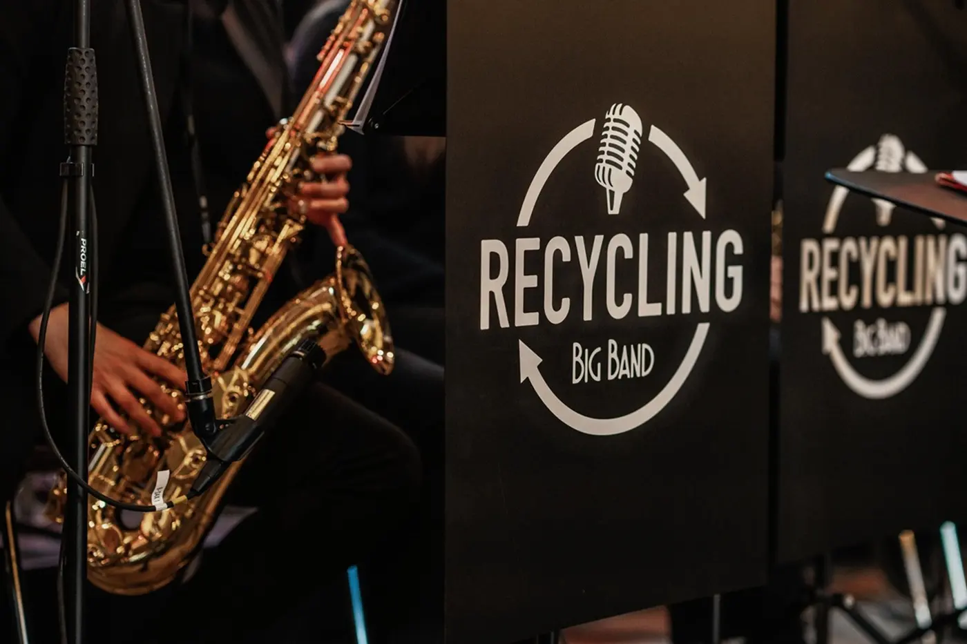 Un sassofonista in abbigliamento scuro mentre suona accanto a un pannello con il logo della band "Recycling Big Band".