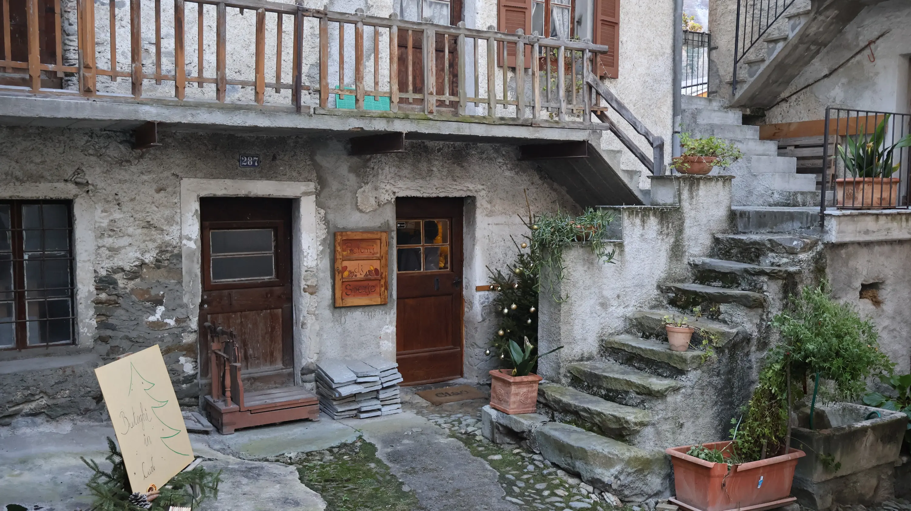 Una piccola corte con due porte, una a sinistra e una a destra, circondata da muri in pietra. Scale di pietra portano a un piano superiore. Vasi