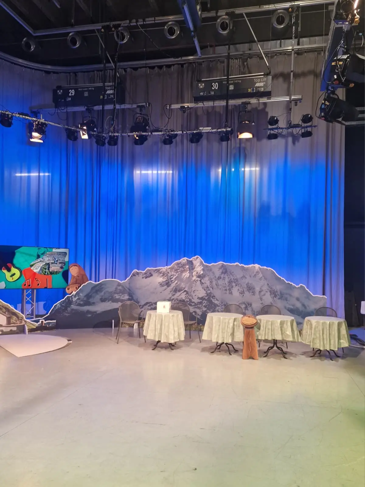 Luce blu dietro a una scenografia di montagne, con tavoli apparecchiati e sedie in uno studio televisivo.