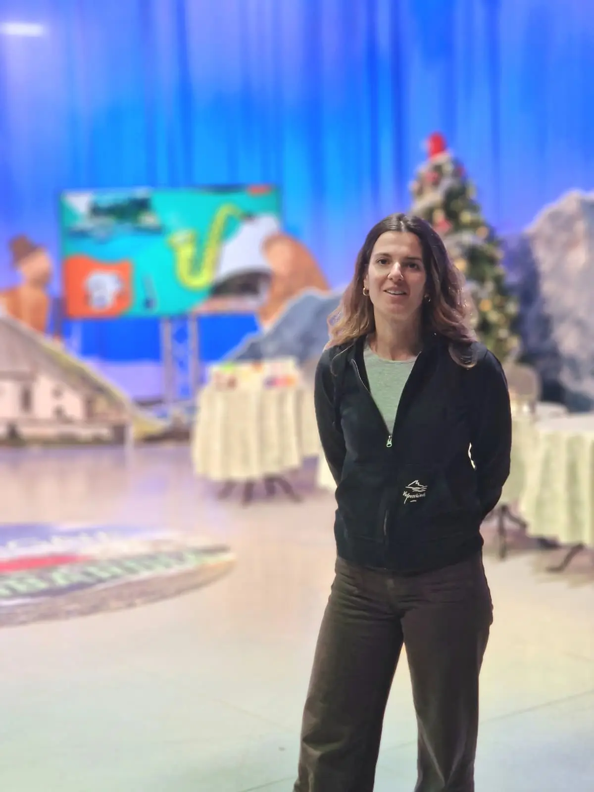 Una donna con i capelli lunghi e mossi indossa un maglione nero e pantaloni marroni, in un ambiente di studio televisivo decorato per le feste, con una grande decor