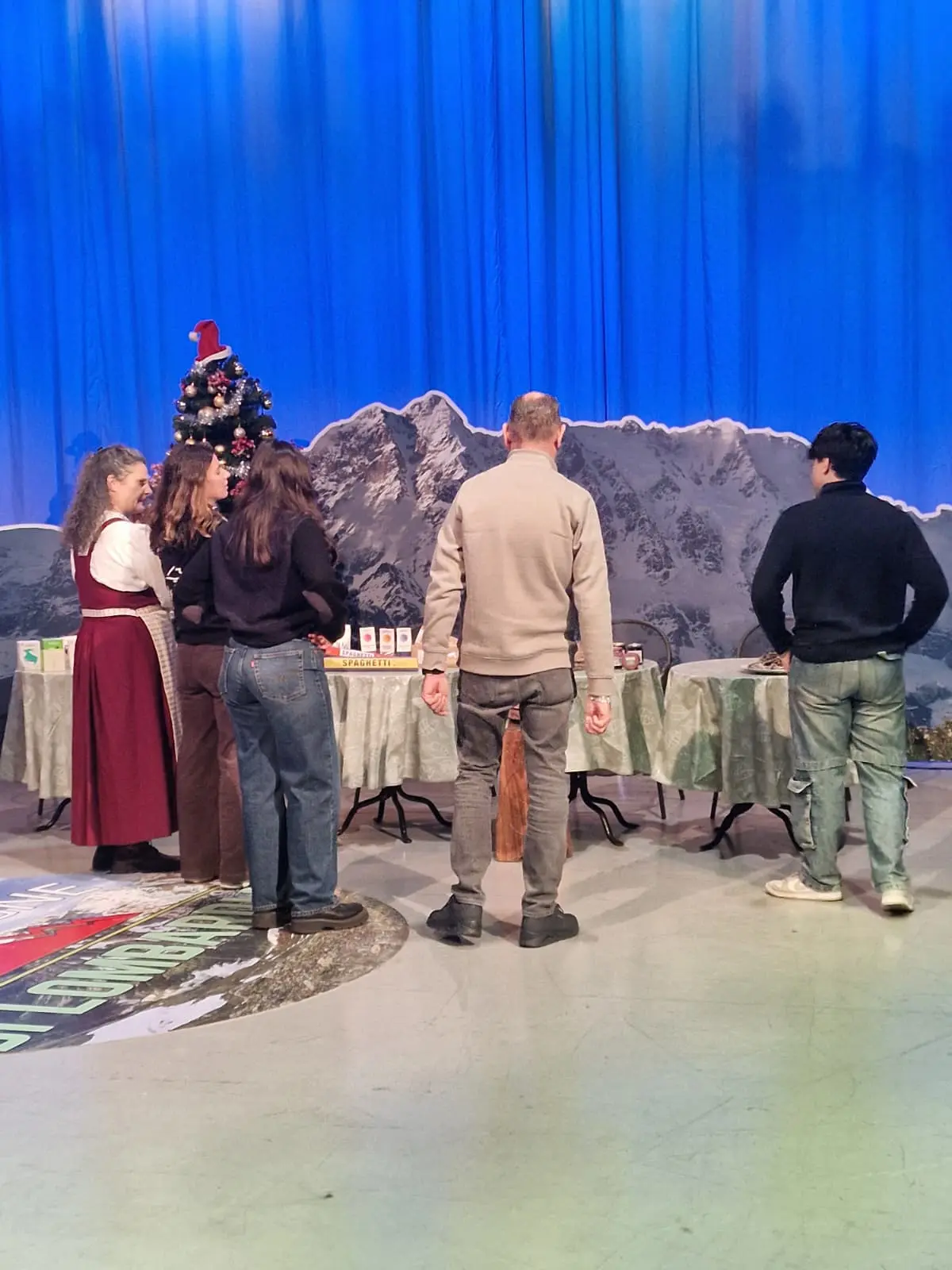 Un gruppo di persone osserva un tavolo allestito con vari oggetti, sullo sfondo una grande montagna. Un albero di Natale decorato si trova accanto al tavolo