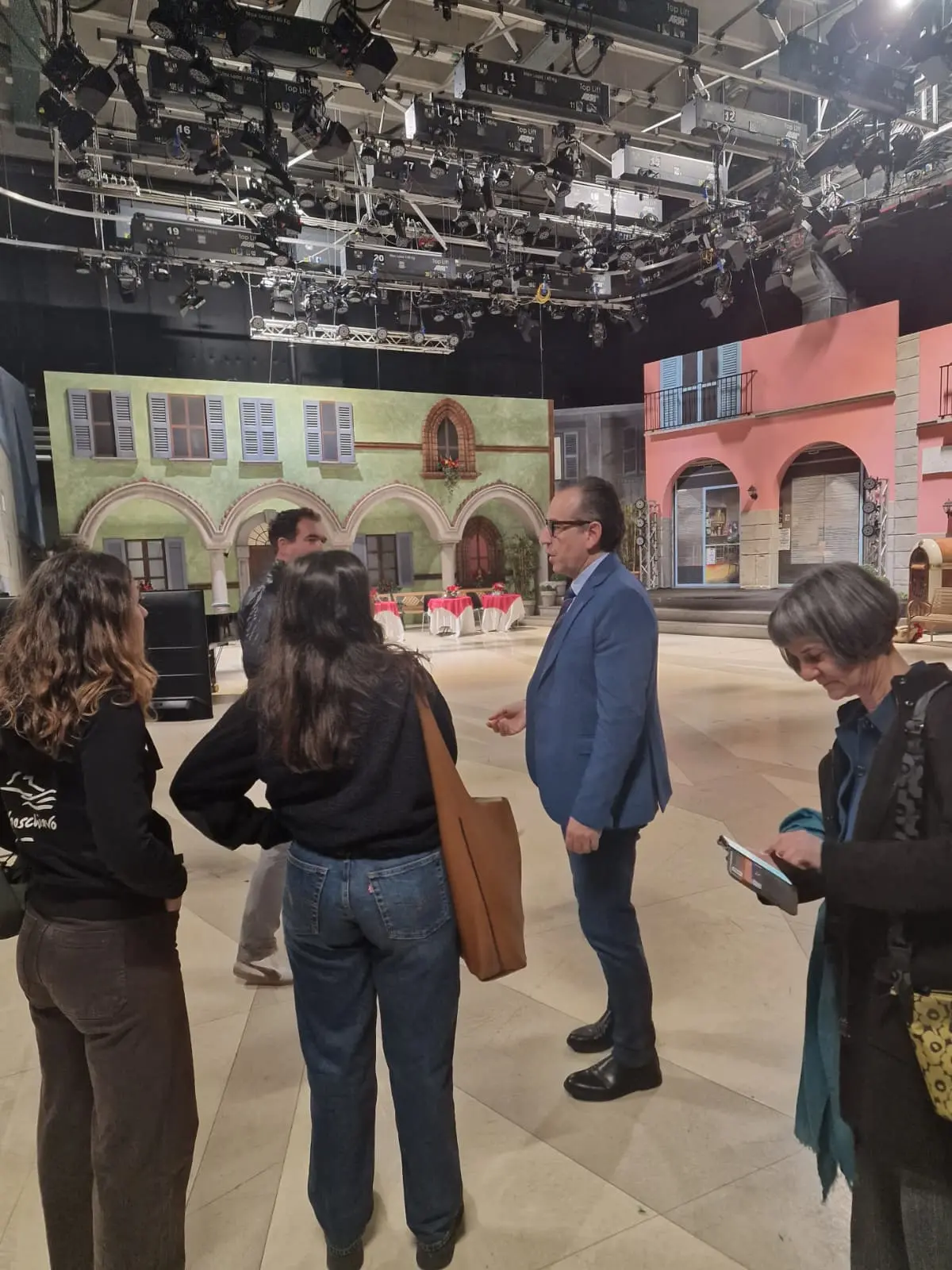 Un gruppo di cinque persone conversano in uno studio televisivo, circondati da scenografie colorate e attrezzature di illuminazione.