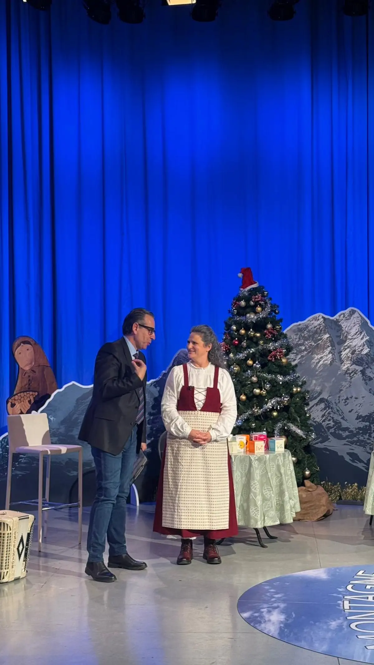 Due persone sul palco di uno spettacolo, una vestita con un grembiule tradizionale e l'altra in abito formale. Sullo sfondo si vede un albero di Natale