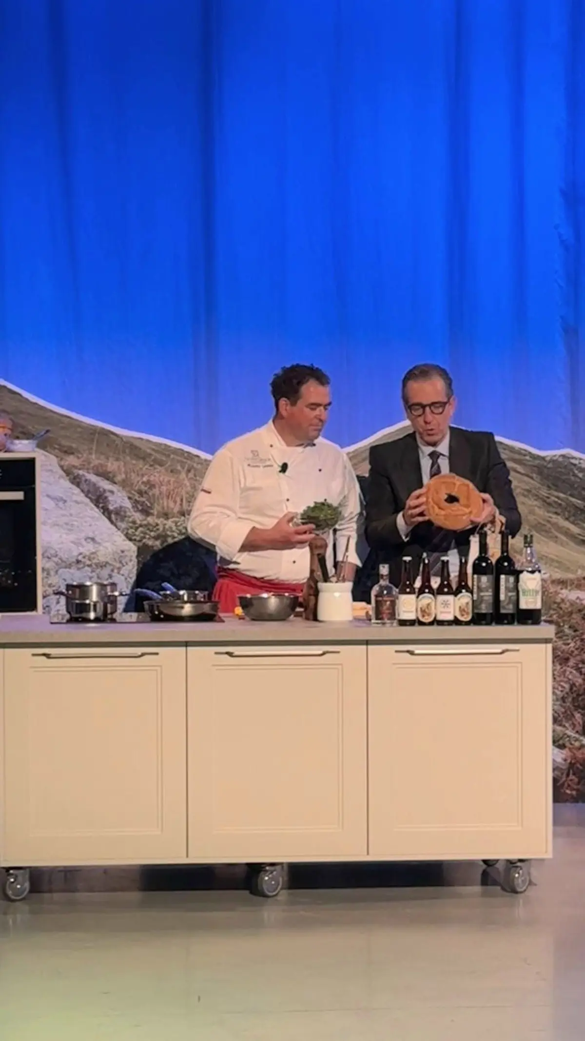 Un cuoco e un presentatore discutono di ingredienti durante una dimostrazione culinaria su un palco. Sul tavolo ci sono pentole, bottiglie e un piatto in legno