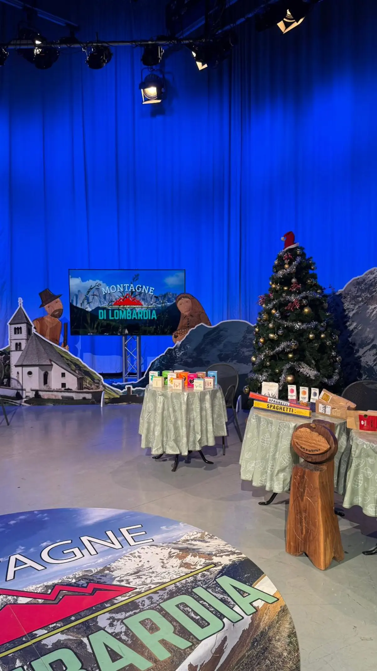 Set di registrazione con decorazioni natalizie, un albero di Natale e tavoli coperti da tovaglie verdi. Sullo sfondo, un video display mostra "Montagne di Lombardia