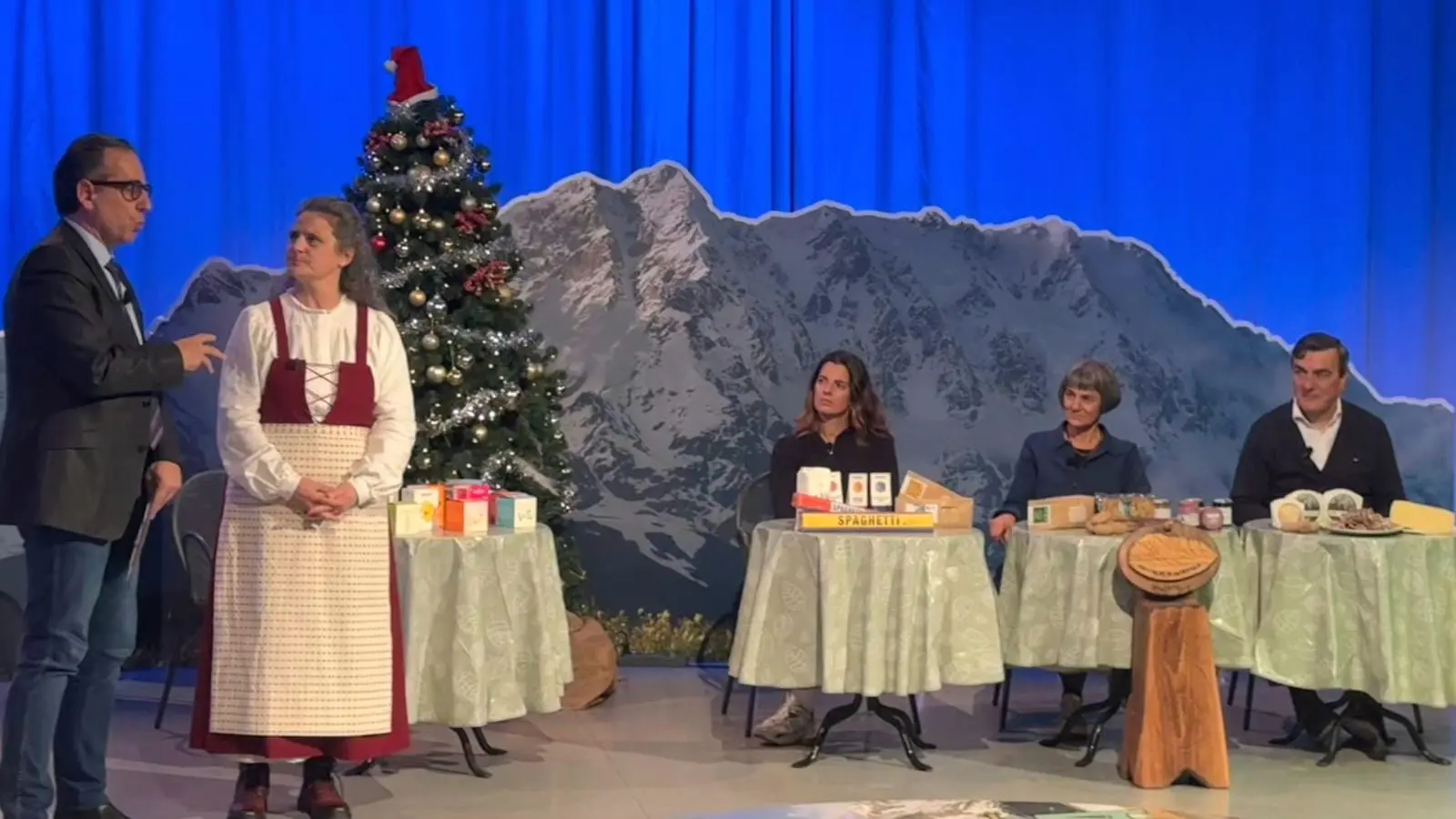 Un set televisivo con un albero di Natale e un paesaggio montano sullo sfondo. Un uomo in giacca parla con una donna vestita con un abito tradizionale