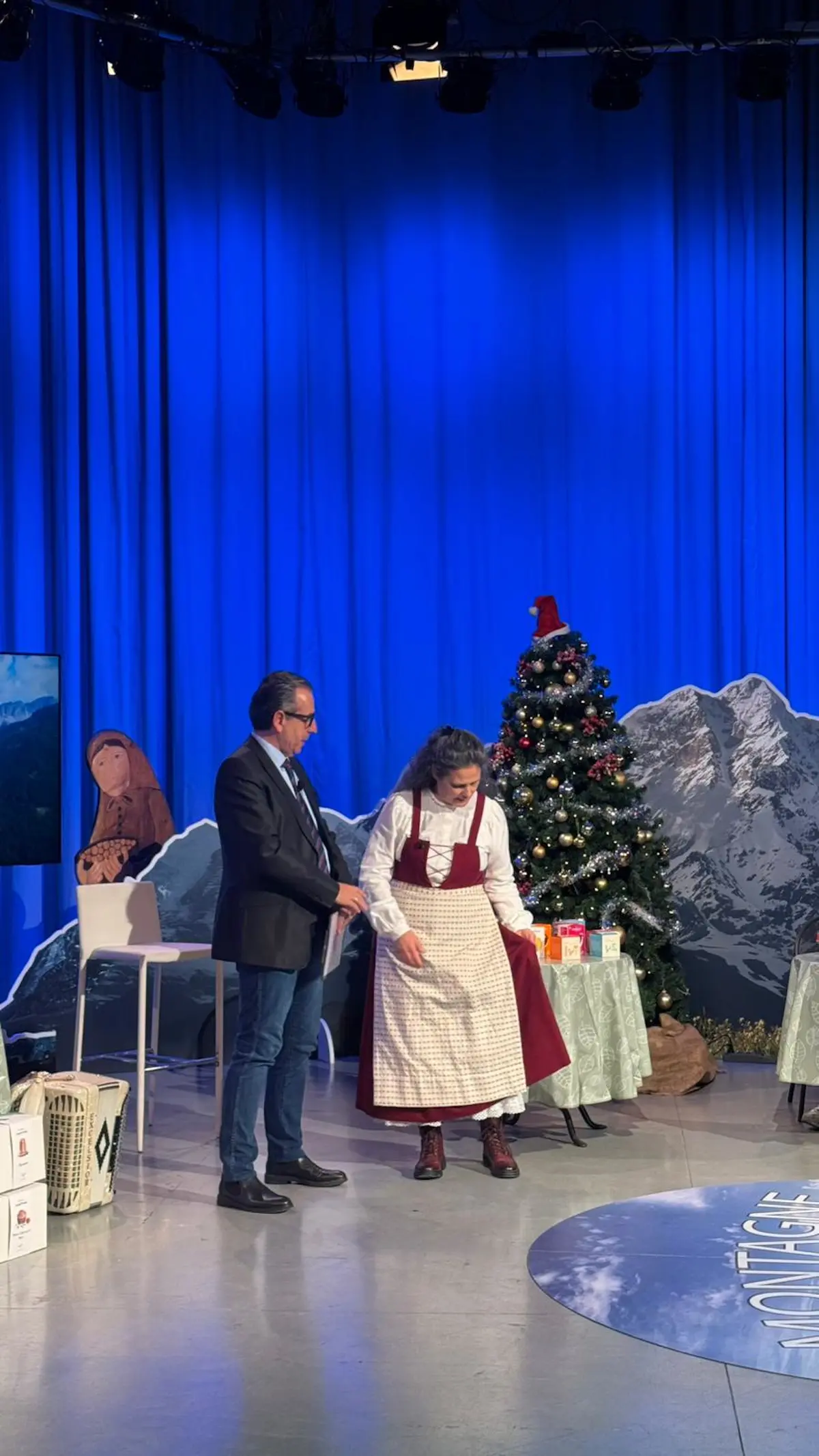 Due persone sono in uno studio televisivo, con un albero di Natale decorato sullo sfondo. Una donna indossa un grembiule e si sta chinando, mentre un uomo in