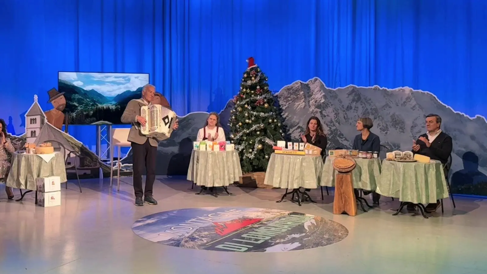 Un uomo suona l'accordeon su un palco decorato con un albero di Natale. Tre donne e due uomini sono seduti a tavoli apparecchiati, mentre un grande sch