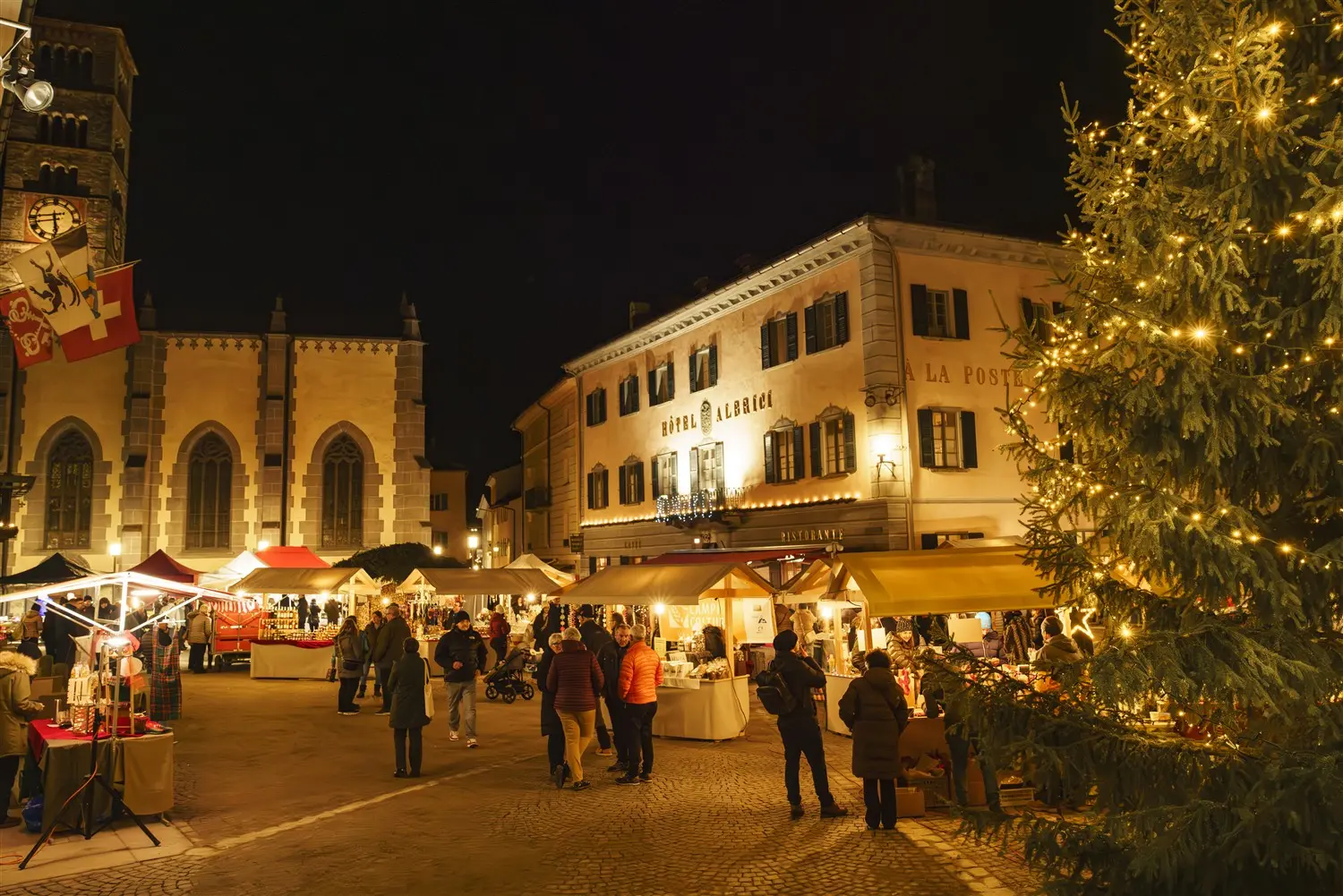 Mercato natalizio in una piazza illuminata, con bancarelle di prodotti artigianali e decorazioni natalizie. Sullo sfondo, un edificio storico e un albero di