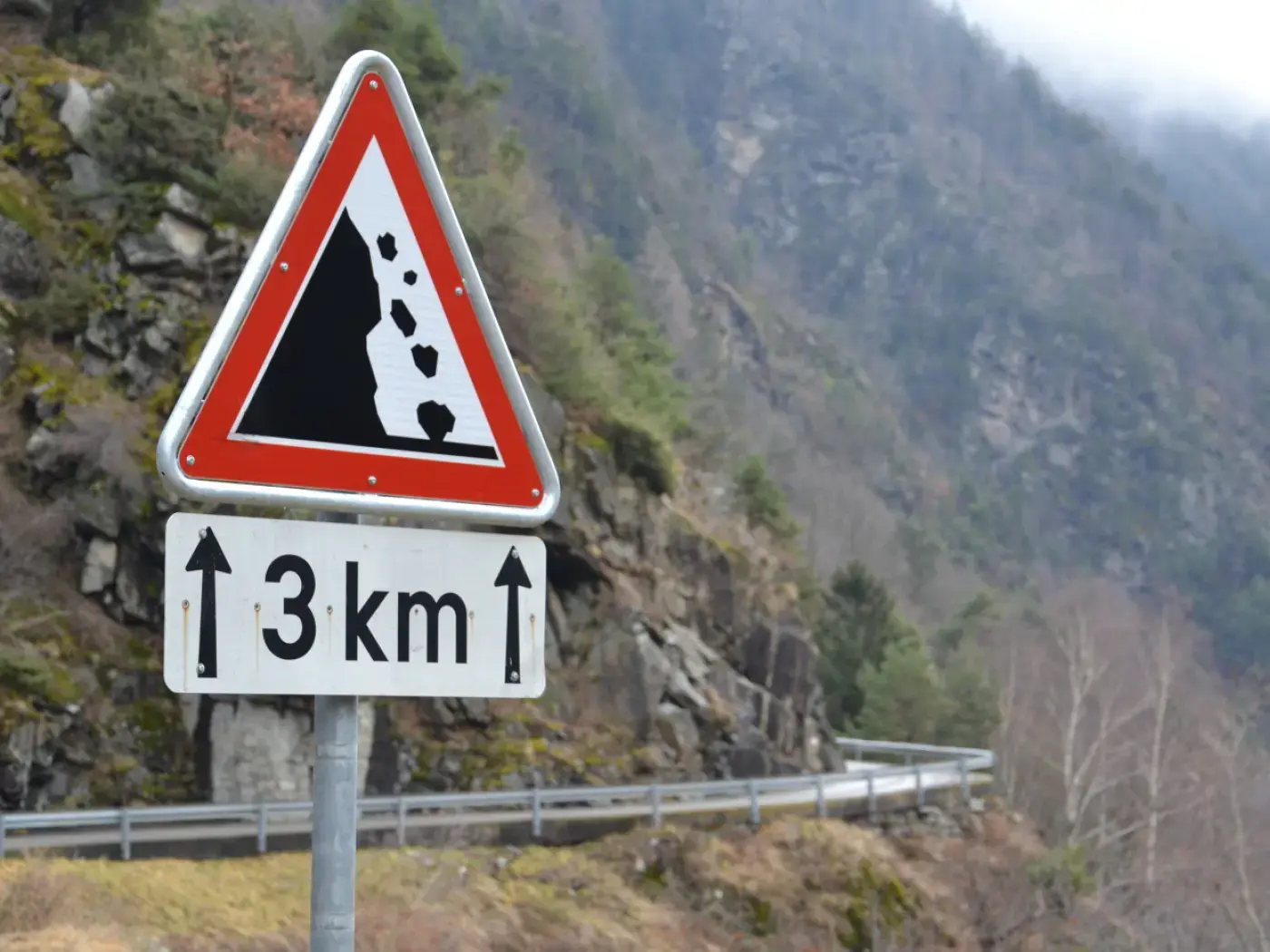 Cartello stradale triangolare che avverte di caduta massi a 3 km, con un'illustrazione di massi che rotolano. Sfondo montano.