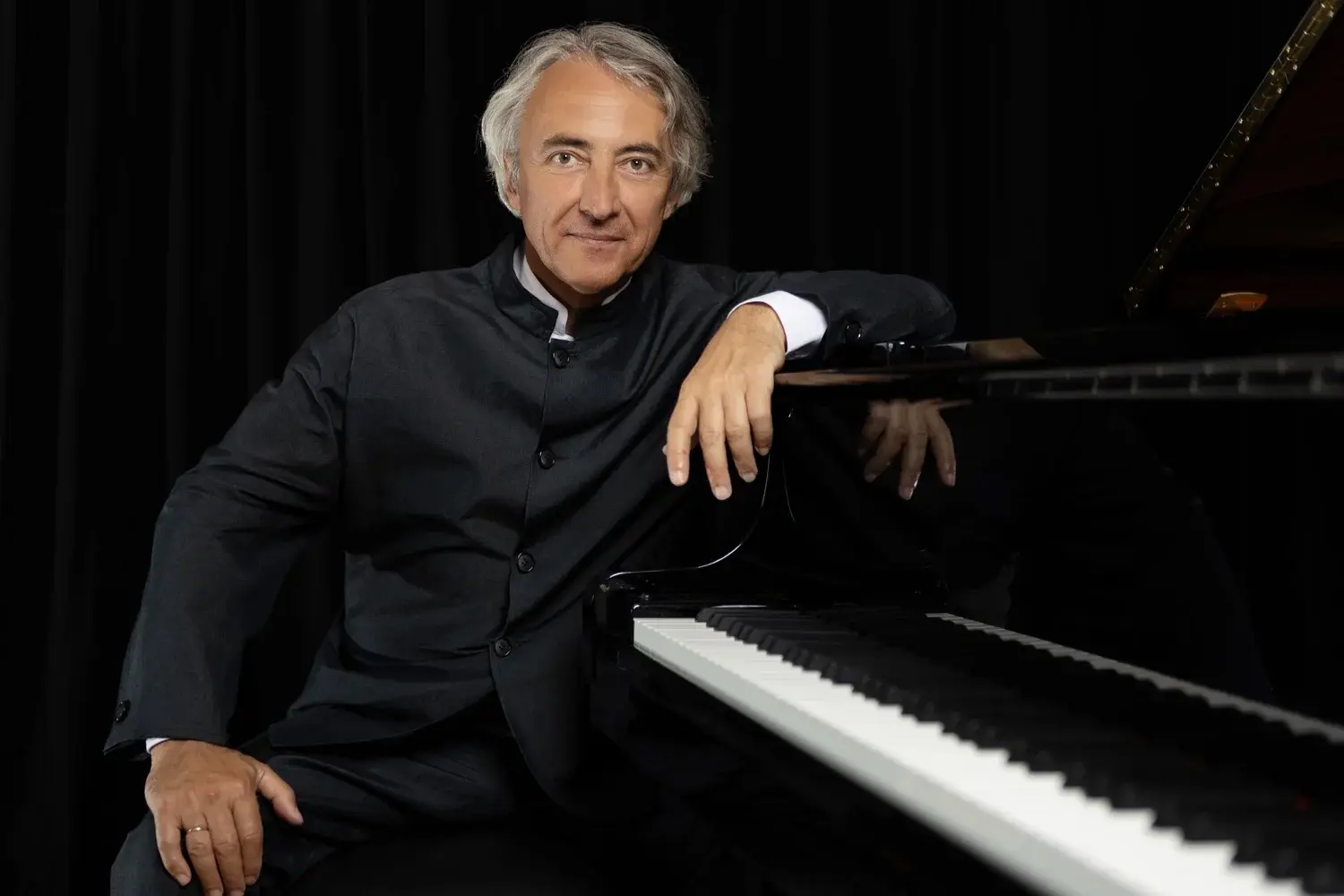 Uomo con capelli grigi e giacca scura, seduto accanto a un pianoforte nero, sorride mentre posiziona una mano sulla tastiera.