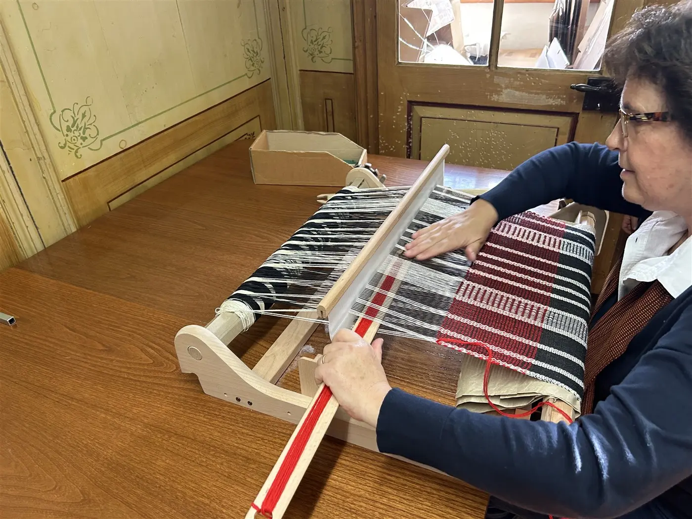 Una donna sta tessendo su un telaio, concentrata mentre sistema i fili. Accanto a lei ci sono diversi materiali e strumenti. La stanza ha pareti di legno e una finestra sullo sfondo.