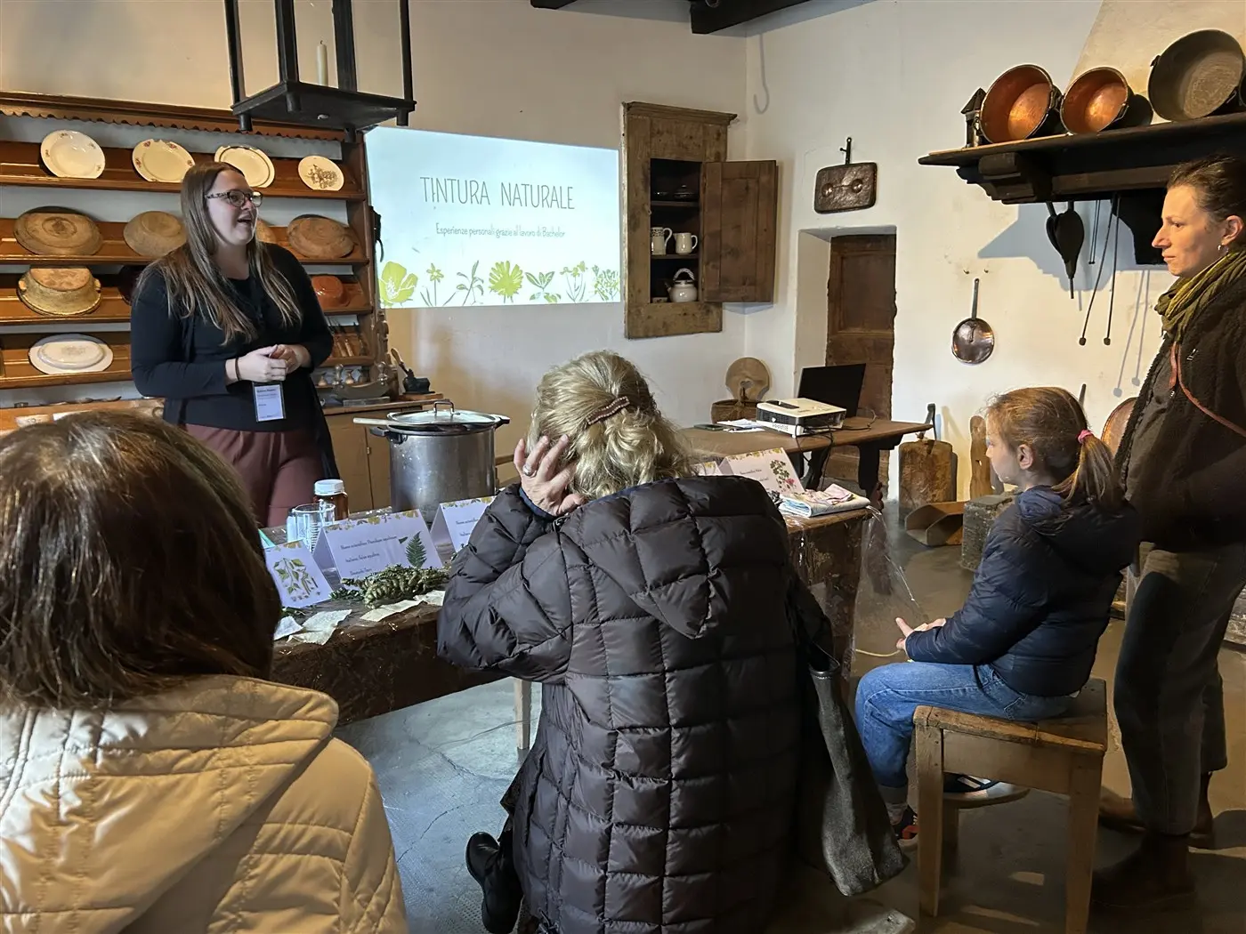 Una donna dà una presentazione sulla tintura naturale a un gruppo di persone sedute. Sul tavolo ci sono materiali per l'attività e sullo sfondo è visibile una proiezione con il titolo della presentazione.